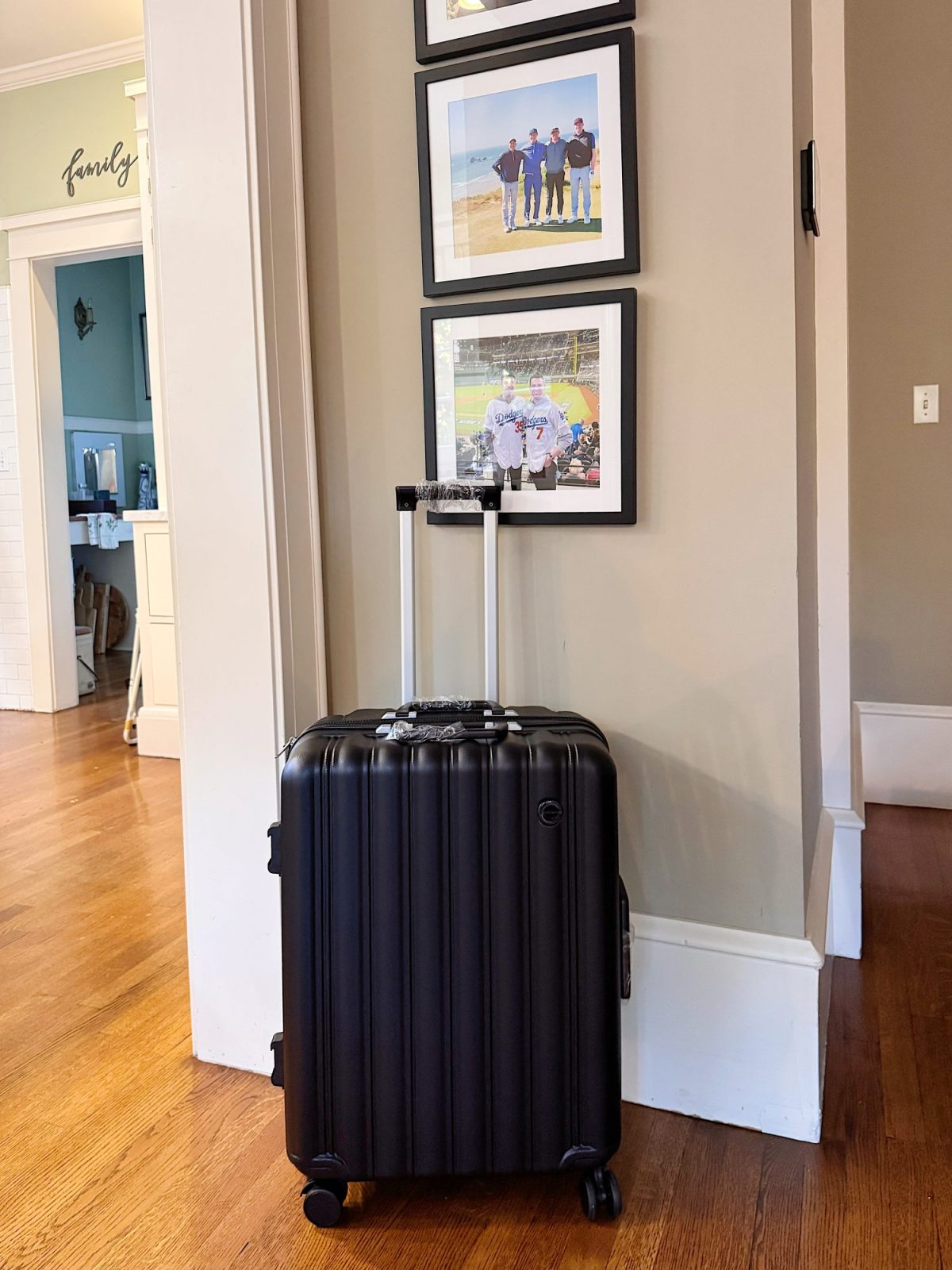 A black hard-shell suitcase with extended handle stands on a wooden floor near a beige wall with framed photos.