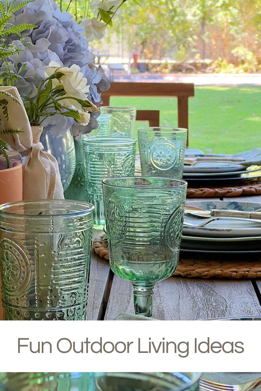 A wooden outdoor table set with green glassware, plates, and floral arrangements, positioned on a lawn with trees in the background.
