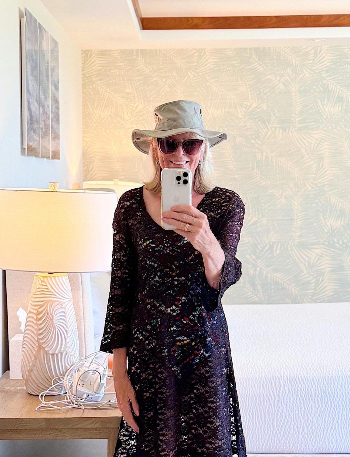 A woman wearing sunglasses, a sun hat, and a black lace dress takes a selfie in a bright, modern bedroom with light-colored decor.