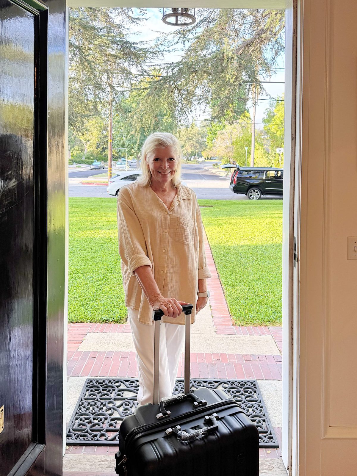 A woman stands at an open front door holding the handle of a black suitcase, with a green lawn and parked cars visible outside.