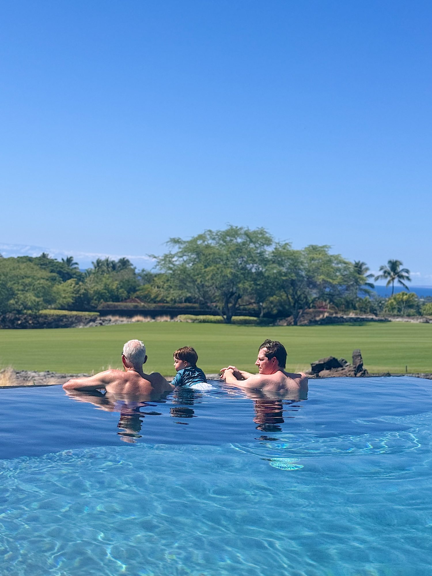 Three people are relaxing at the edge of an infinity pool, facing a green lawn with trees and a clear blue sky in the background.
