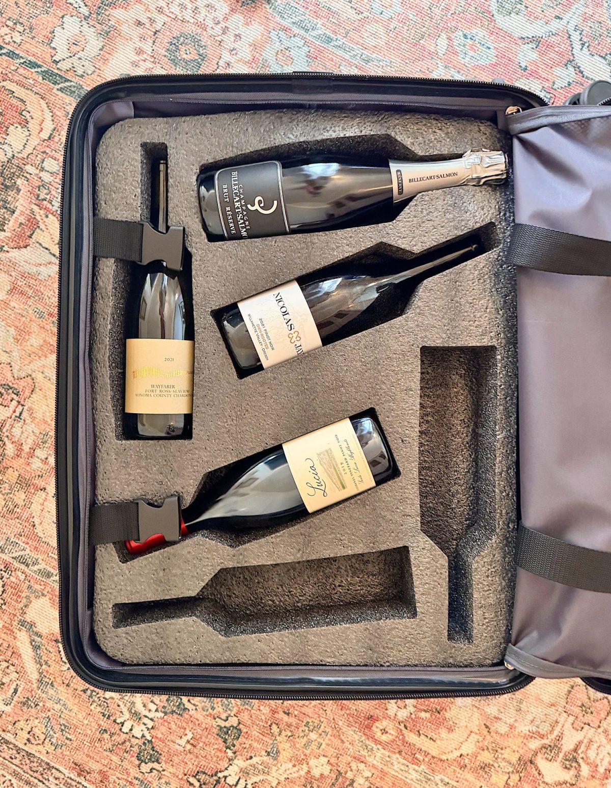 An open suitcase with foam inserts securely holding four bottles of wine, placed on a patterned rug.