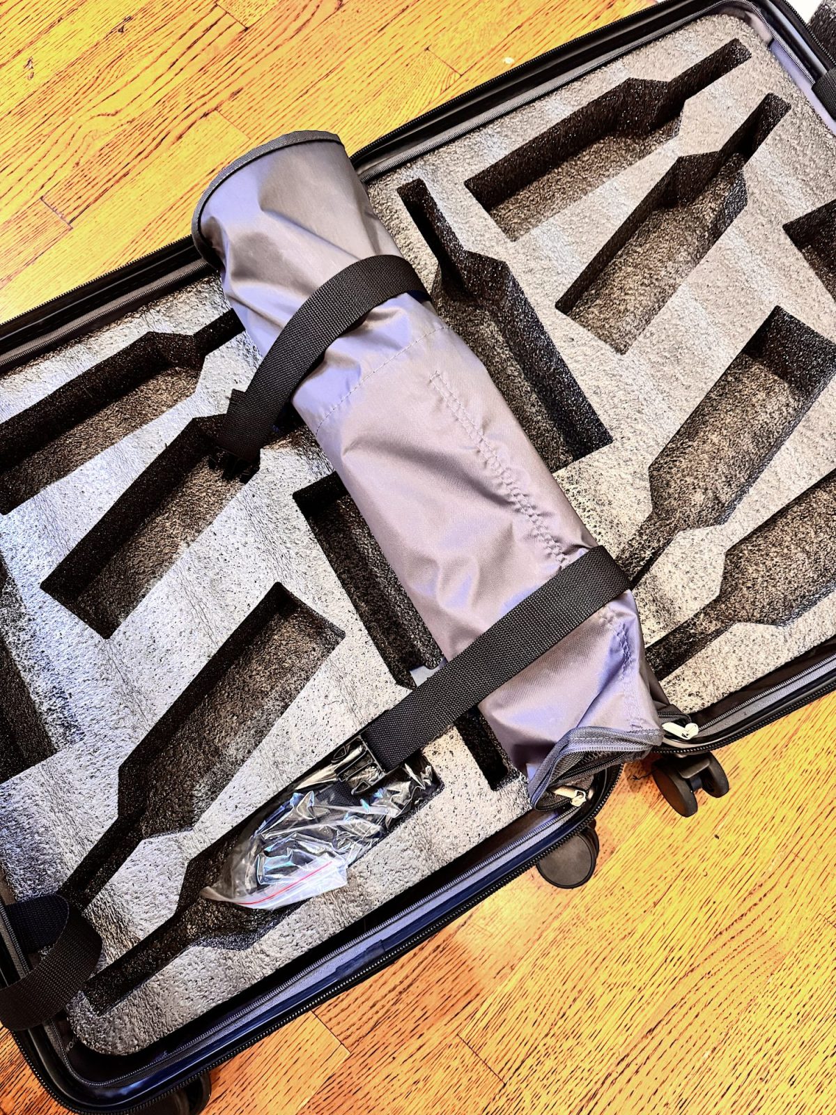 A padded hard case with custom foam inserts holds a cylindrical object wrapped in a gray protective sleeve secured by two straps. The case is open on a wooden floor.