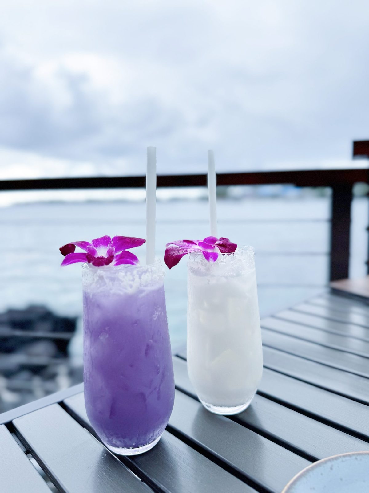 Two tropical drinks, one purple and one white, each garnished with a purple orchid, sit on a table overlooking the ocean.