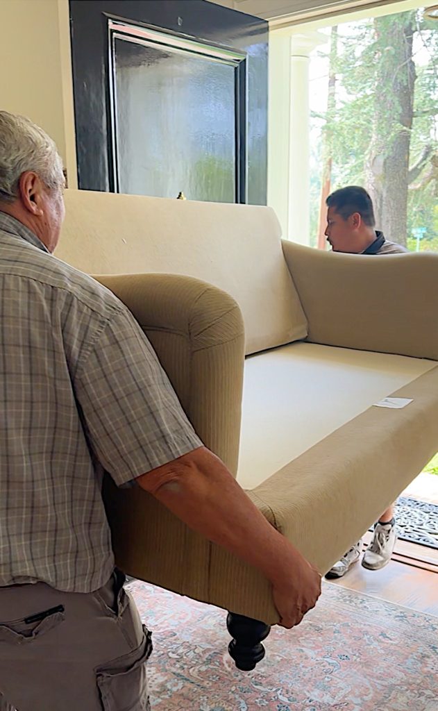 Two people are carrying a beige upholstered sofa through a doorway into a house.