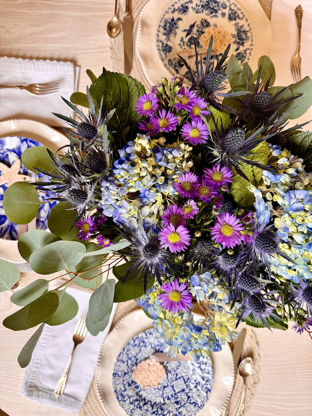 A bouquet of purple, blue, and green flowers is arranged on a dining table set with blue and white plates, beige placemats, and gold-colored utensils.