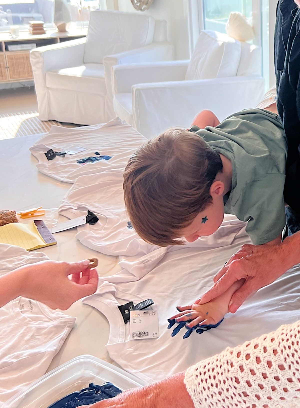 A child is pressing a blue paint-covered hand onto a white T-shirt with the help of an adult; several blank T-shirts and supplies are on the table.