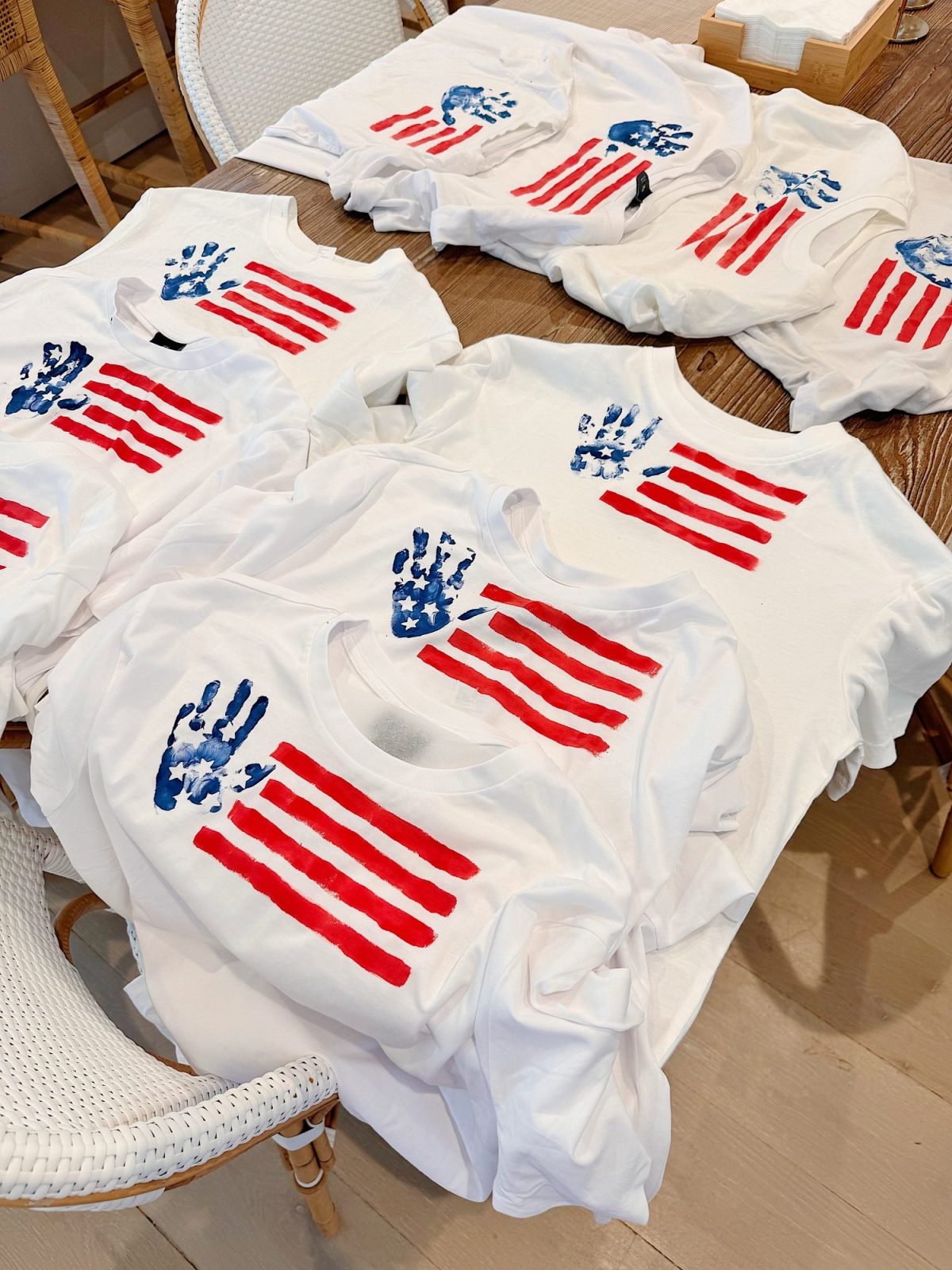 White t-shirts with painted American flag designs, featuring red stripes and blue handprints for the stars, are laid out on a table.