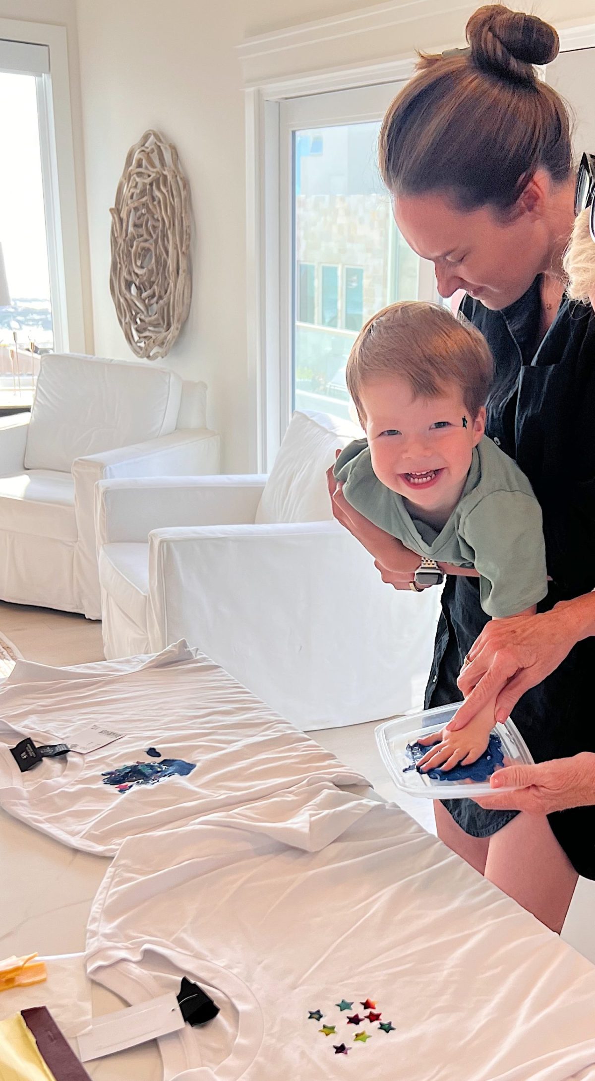 A woman helps a smiling young boy apply paint to a t-shirt on a table in a bright, modern room.