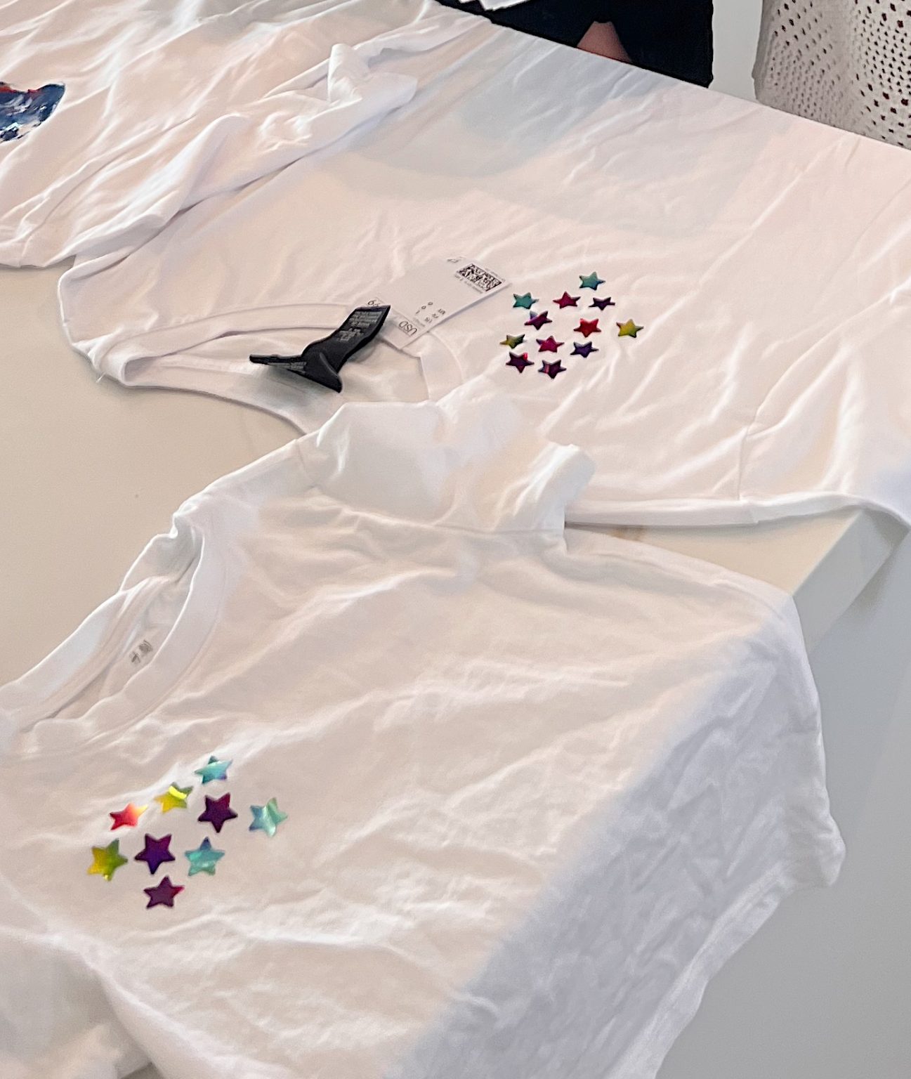 Three white t-shirts are laid out on a table, each decorated with colorful star-shaped stickers on the front.