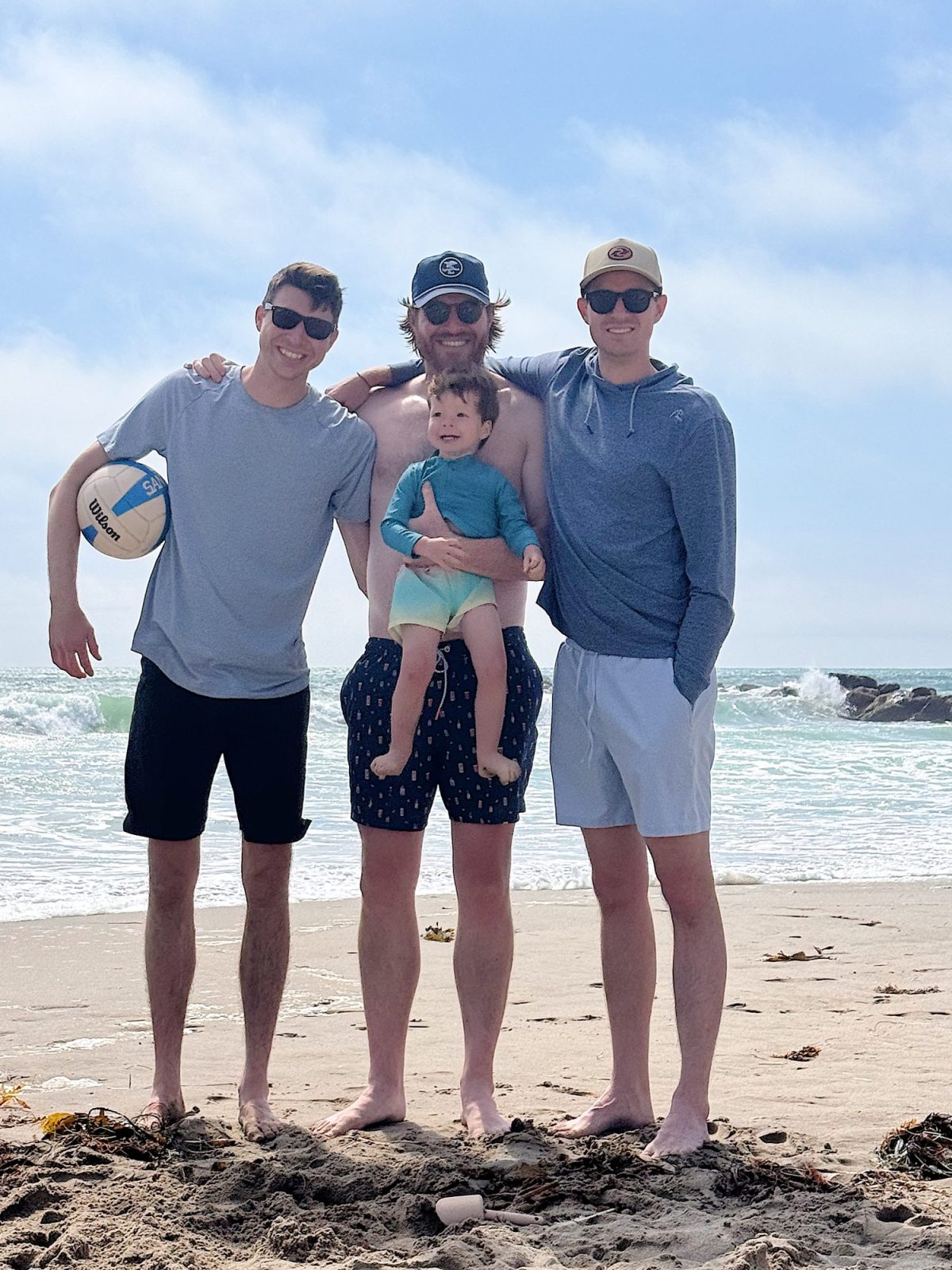 Three men and a small child stand on a sandy beach near the ocean; one man holds a volleyball, another holds the child, and waves are visible in the background.