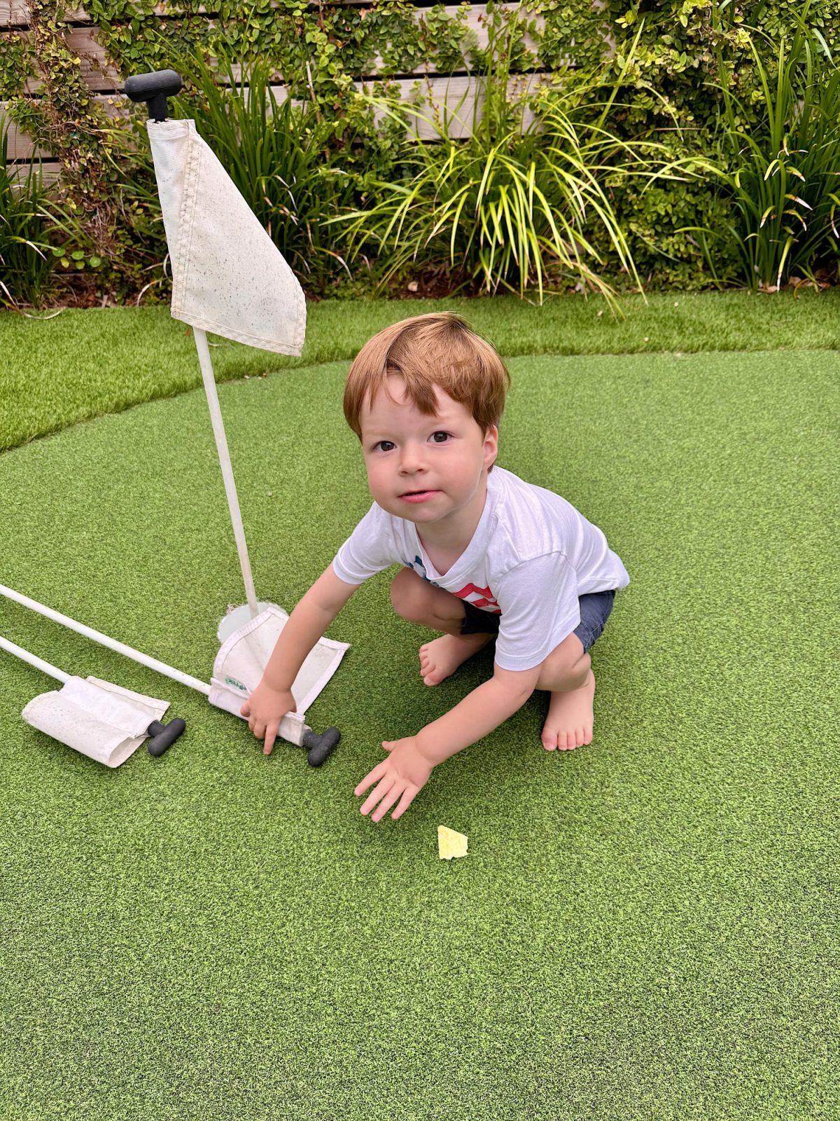 A young boy in a white t-shirt and shorts crouches barefoot on artificial grass, holding a mini golf flagpole. There are two flagpoles and some greenery in the background.