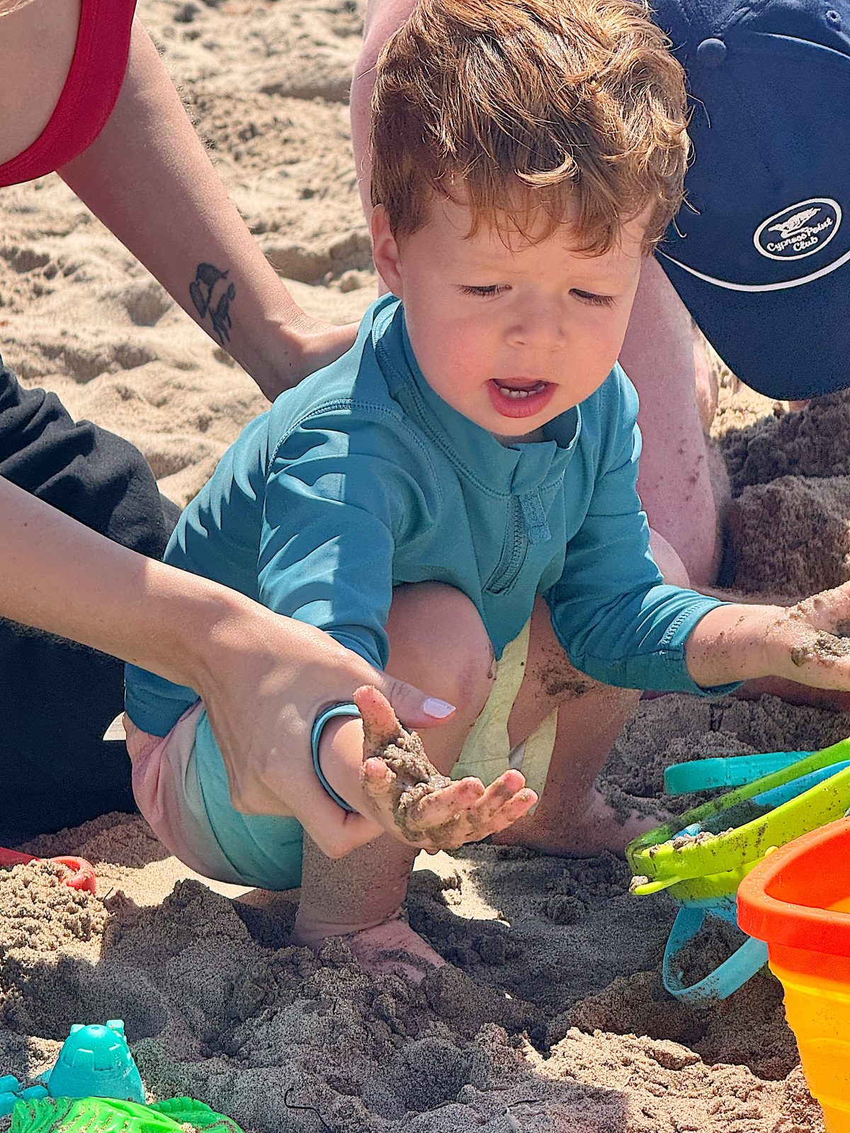 A young child in a blue rash guard sits in the sand at the beach, examining sand on his hands while an adult assists. Colorful sand toys are nearby.