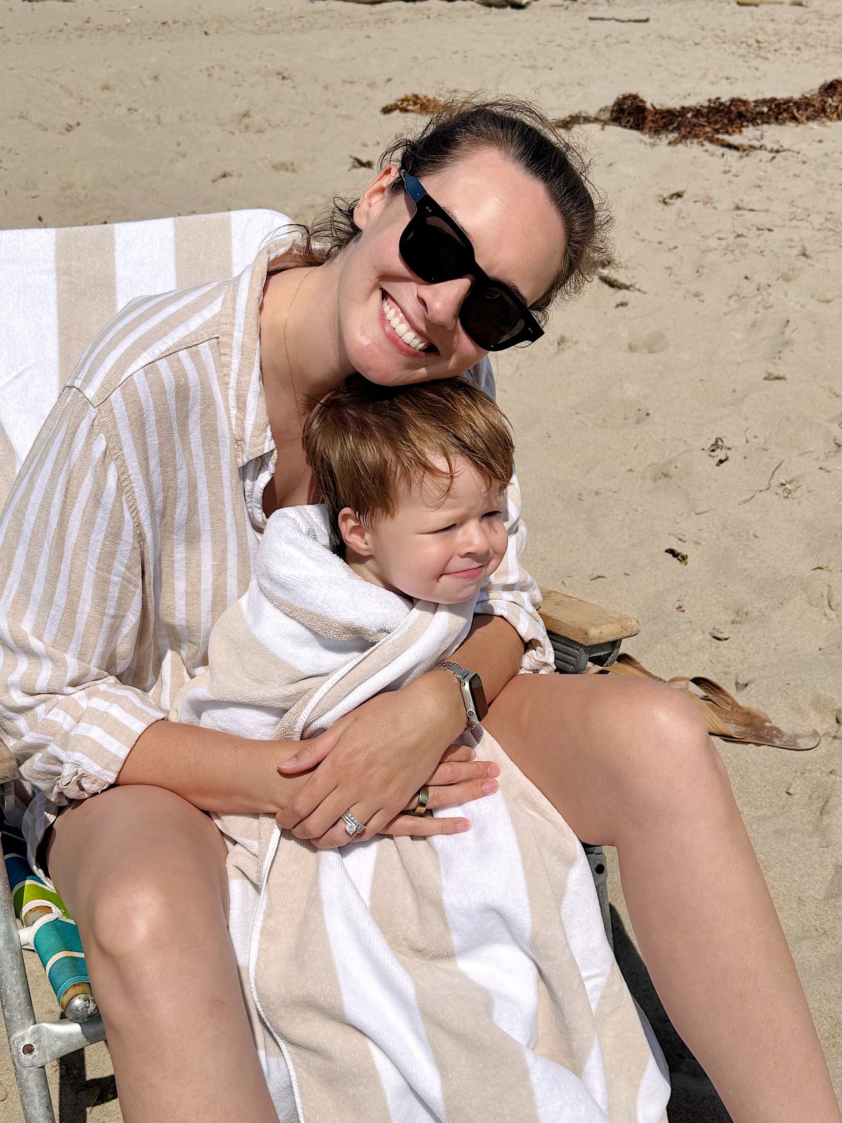 An adult woman in sunglasses hugs a young child wrapped in a towel while sitting on a beach chair at the sandy beach.