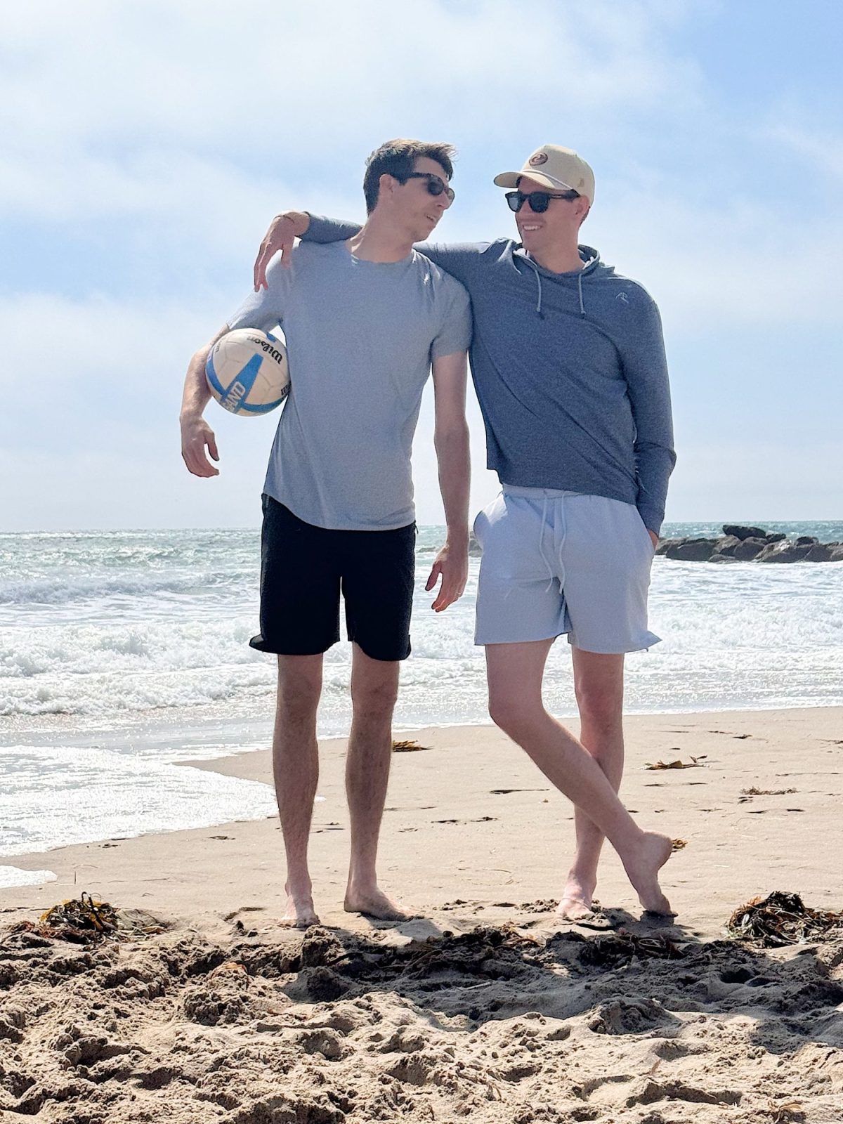 Two men stand barefoot on a sandy beach, one holding a volleyball. Both are wearing sunglasses and casual clothing, smiling at each other with the ocean in the background.