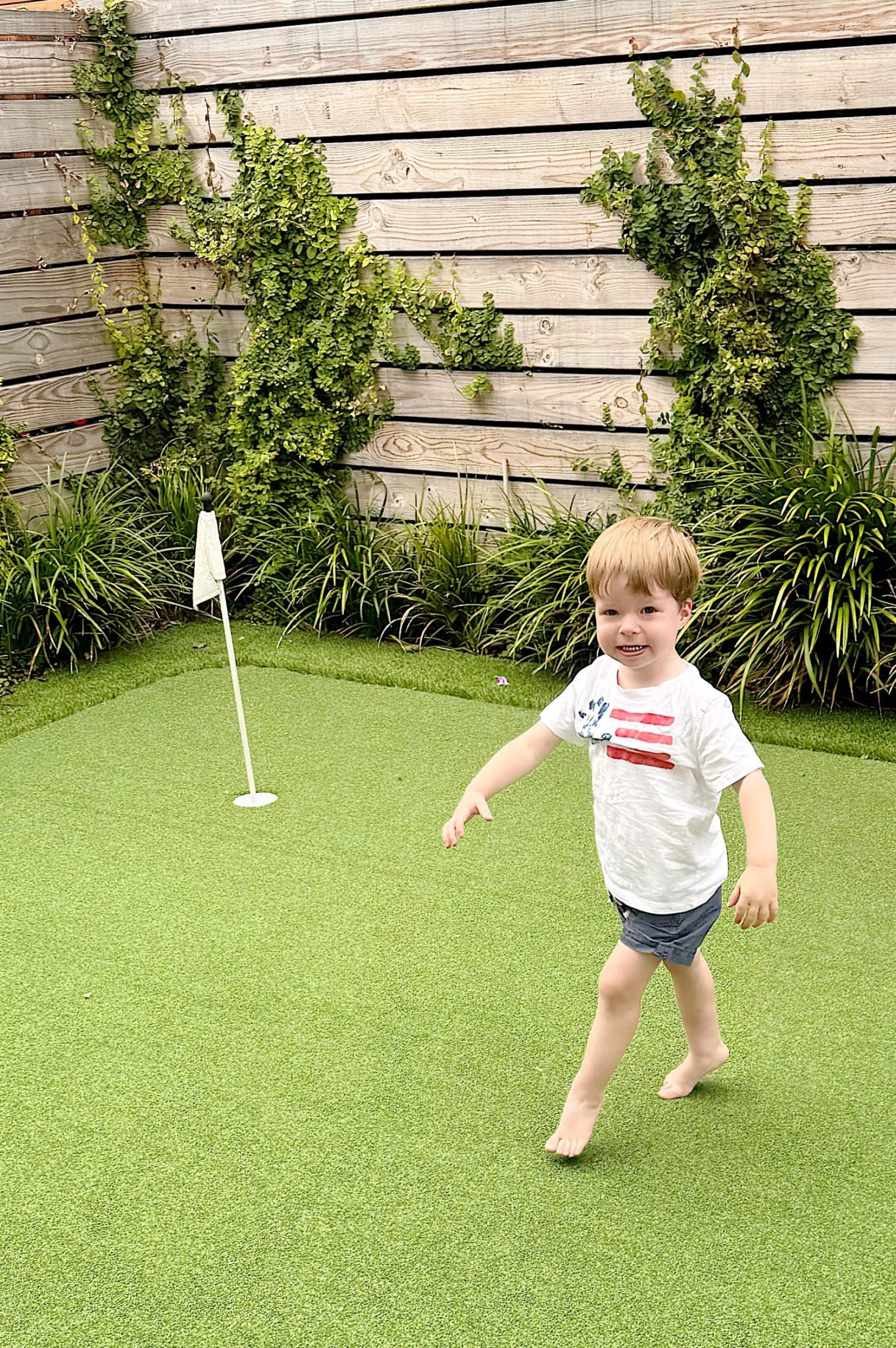 A young boy walks barefoot on artificial grass near a small golf putting flag, with wooden fencing and green plants in the background.