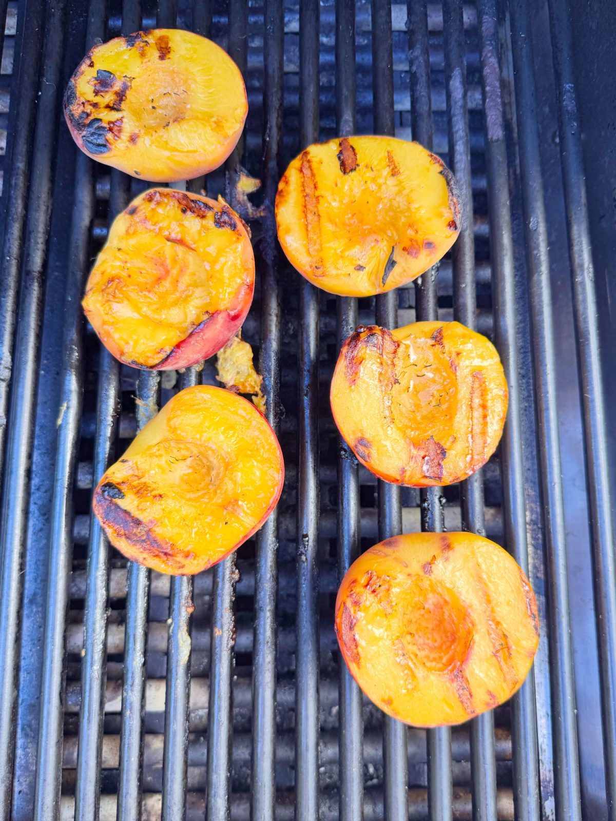 Six peach halves are being grilled on a barbecue, with visible grill marks and slight charring on the fruit.