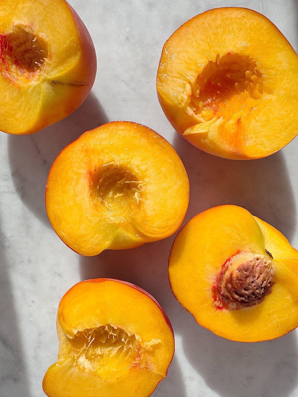 Five halved peaches with pits removed are arranged on a white surface, with sunlight casting shadows across the scene.