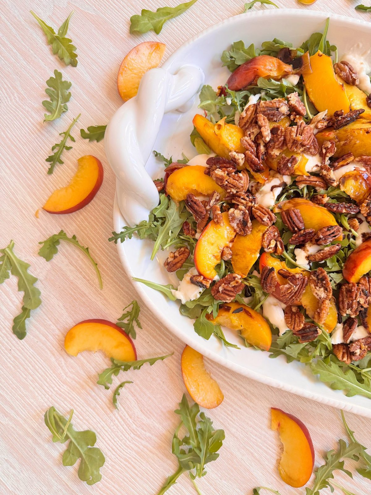 A salad with arugula, sliced peaches, pecans, and mozzarella sits on a white plate, with extra arugula and peach slices scattered on a light wooden surface.