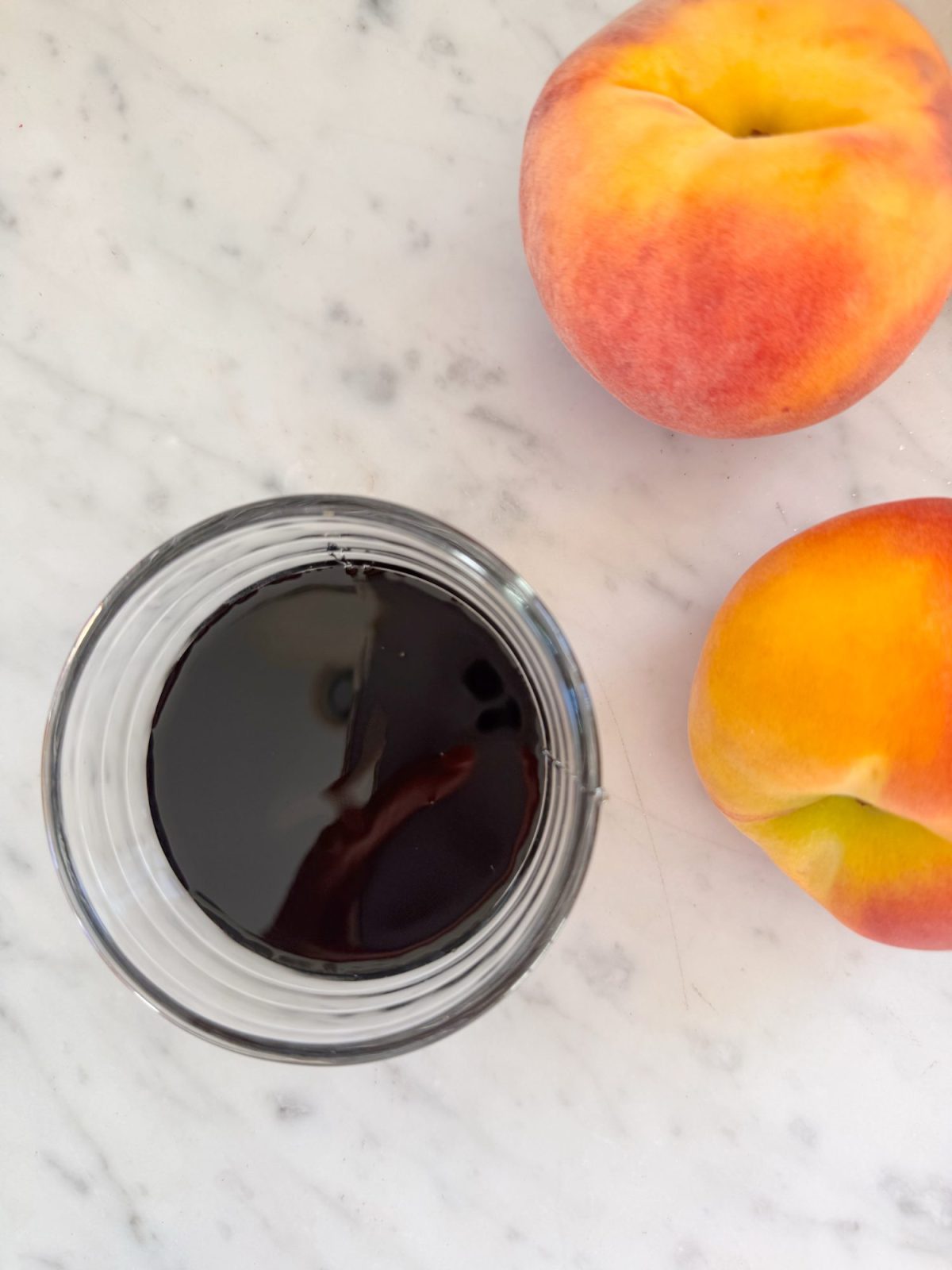 A glass jar filled with dark liquid sits on a marble surface next to two ripe peaches.