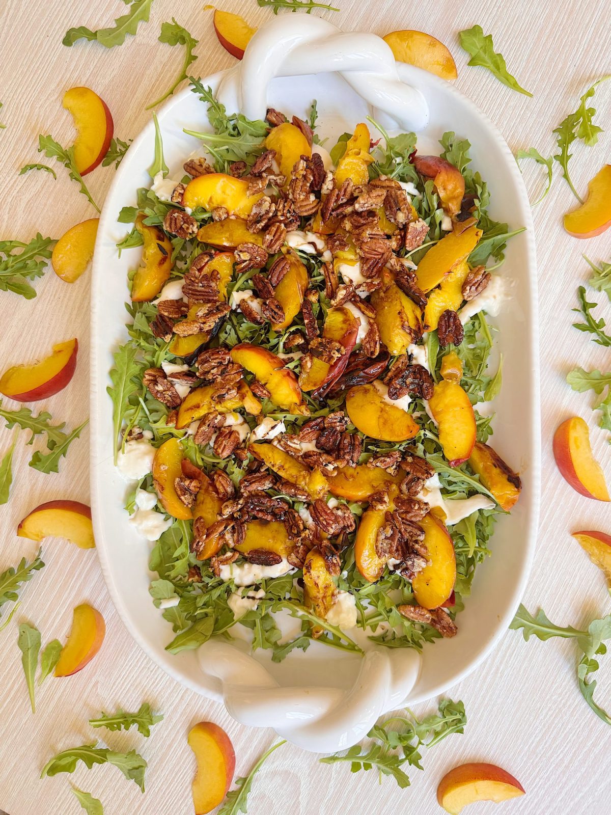 A serving platter of arugula topped with sliced peaches, candied pecans, and a creamy white cheese, garnished with extra arugula and peach slices around the dish.