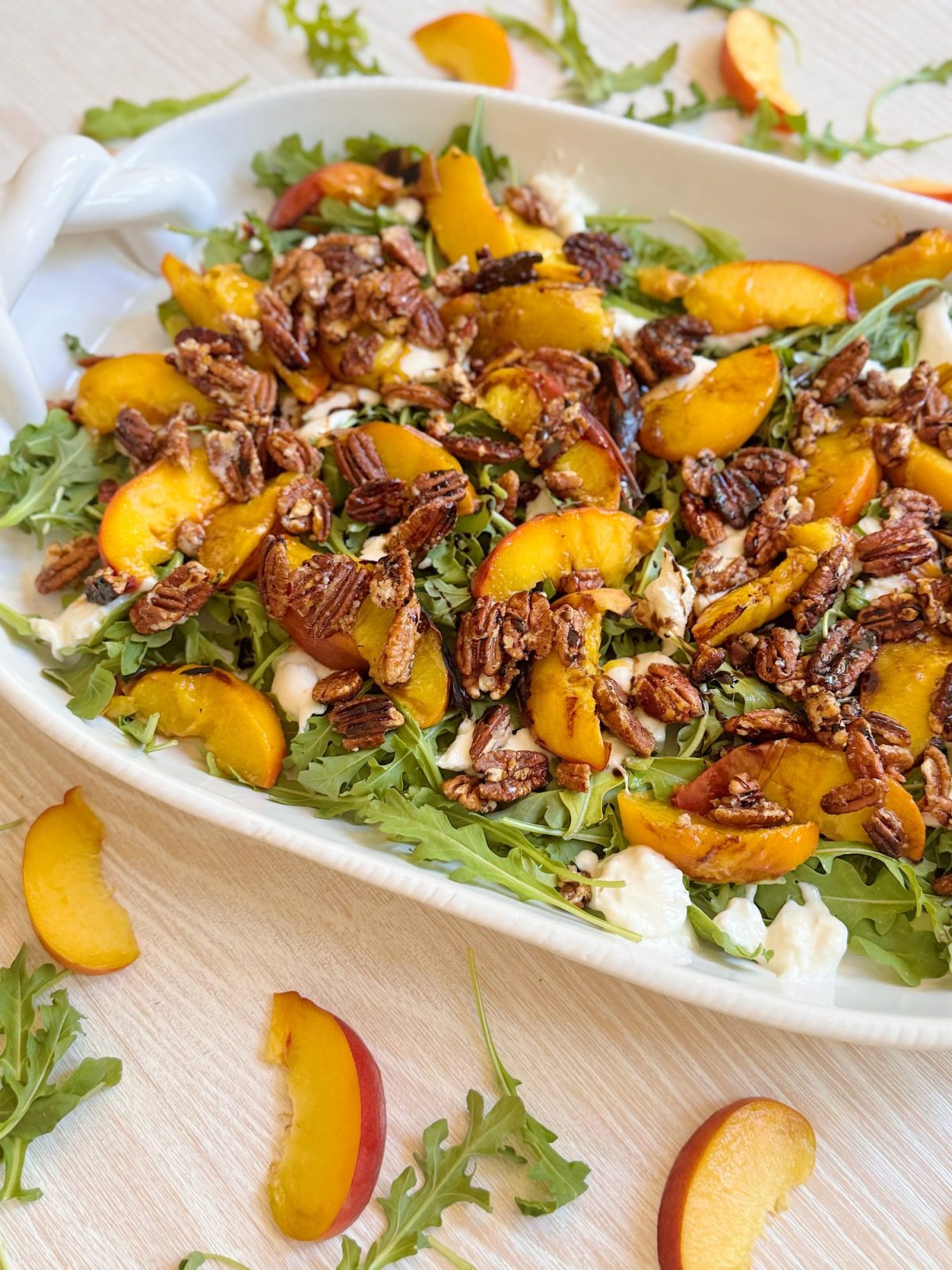 A salad with arugula, sliced peaches, candied pecans, and pieces of white cheese is served in a white dish, with extra peach slices and arugula scattered on the table.