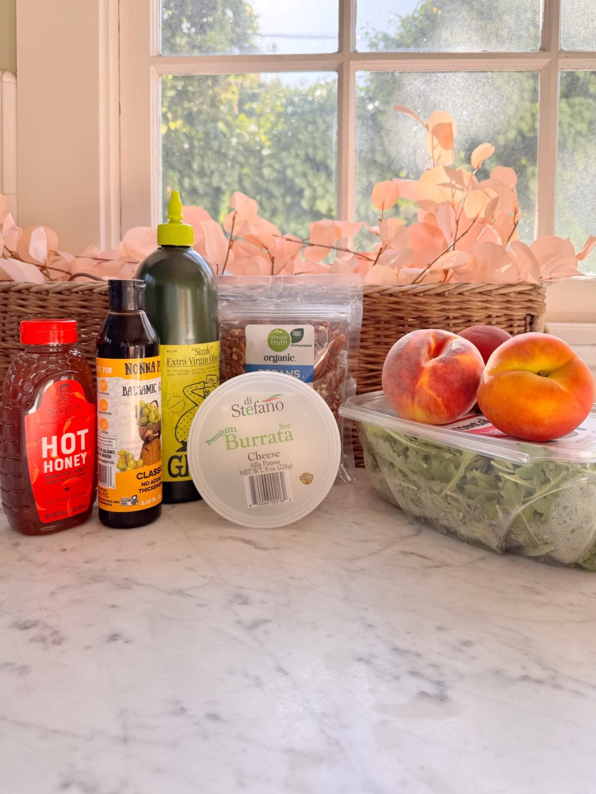 Various food items including hot honey, balsamic glaze, olive oil, pecans, peaches, burrata cheese, and arugula are arranged on a marble countertop in front of a window.