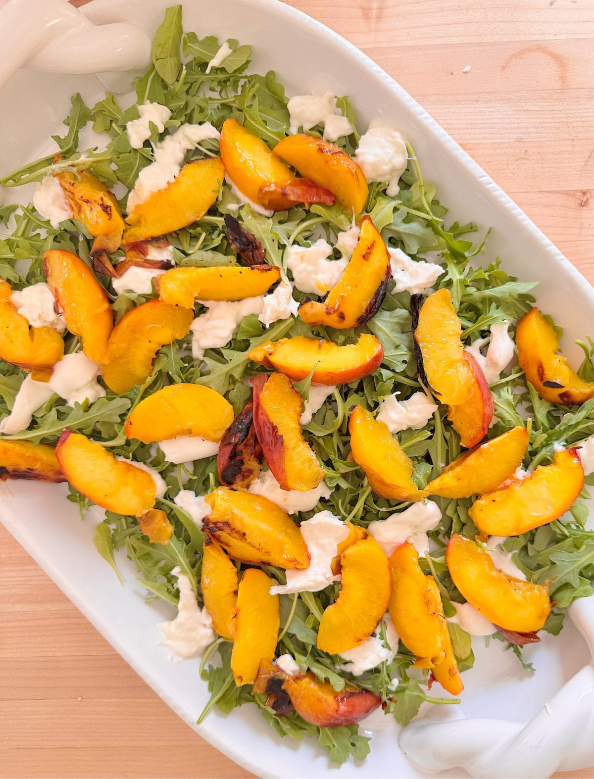 A white dish filled with arugula, grilled peach slices, and dollops of fresh mozzarella cheese on a wooden surface.