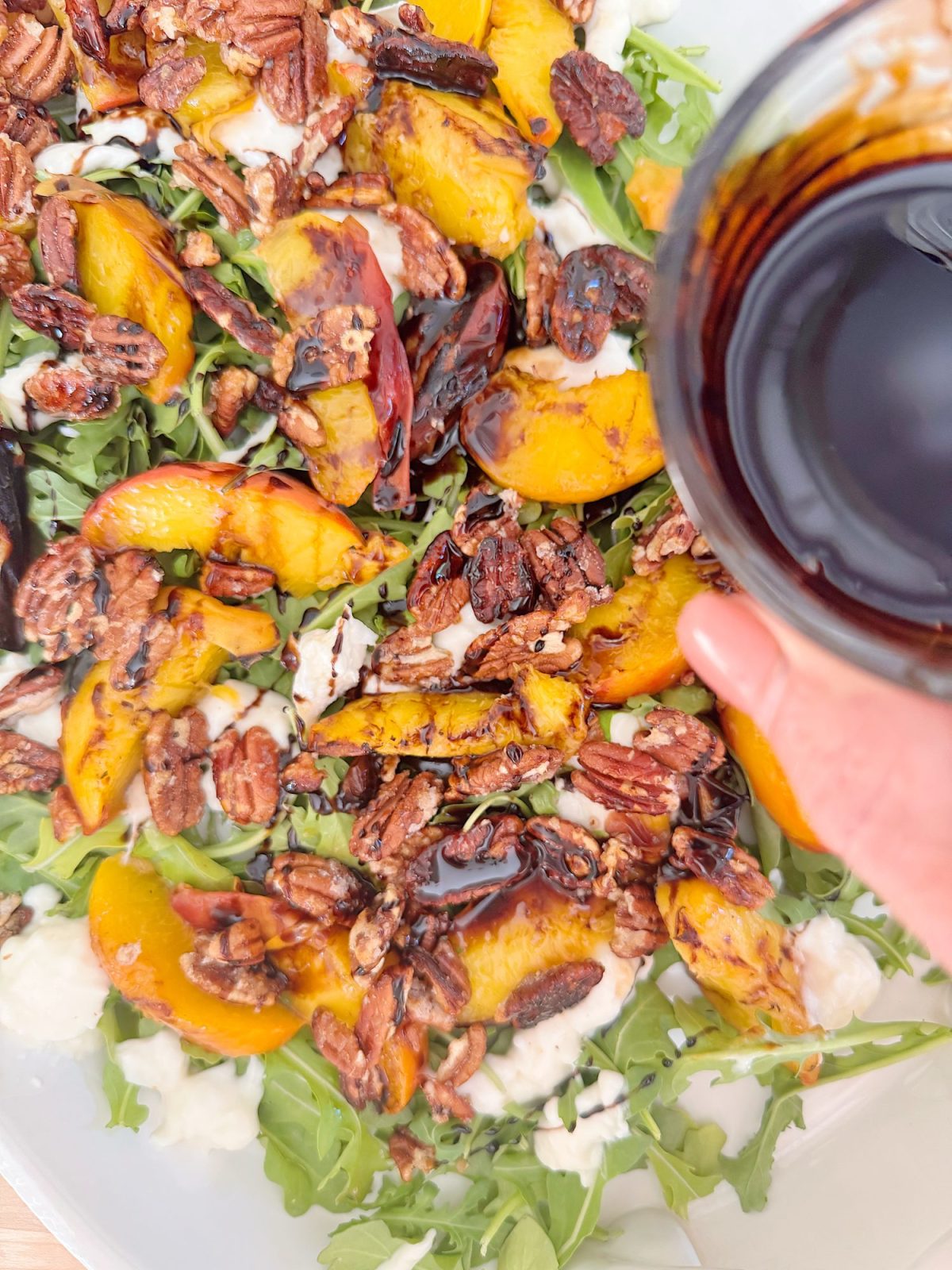 A hand pours balsamic glaze onto a salad with arugula, grilled peaches, pecans, and crumbled cheese in a white bowl.
