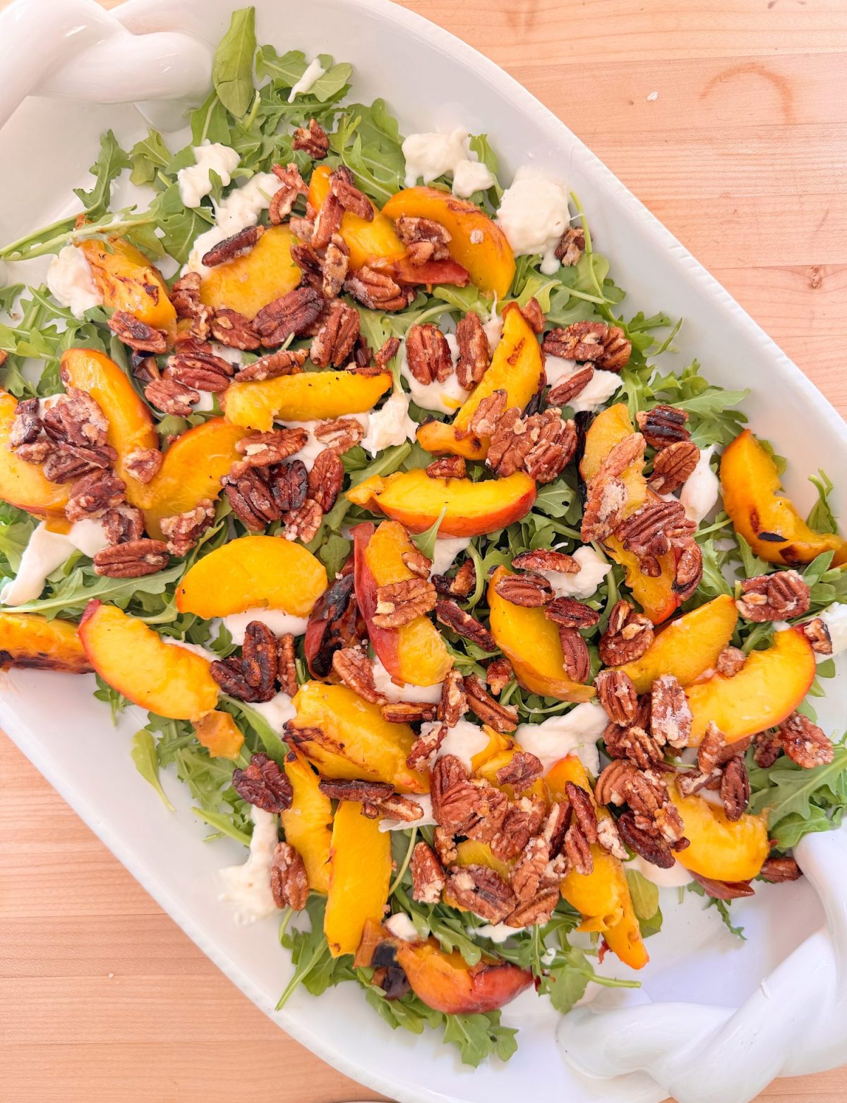 A salad with arugula, sliced peaches, candied pecans, and pieces of cheese arranged on a white serving dish.