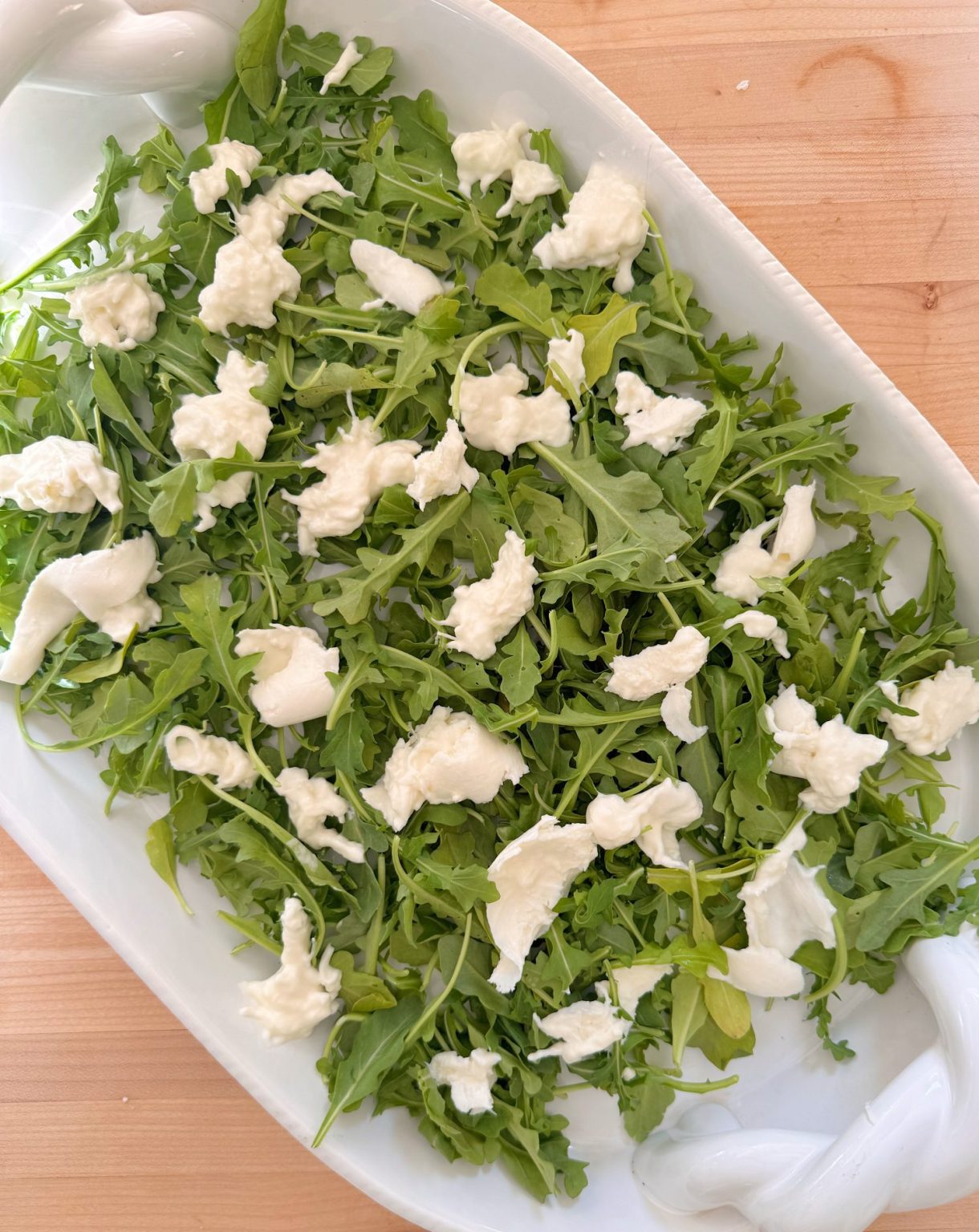 A white dish filled with fresh arugula leaves topped with chunks of mozzarella cheese, placed on a wooden surface.