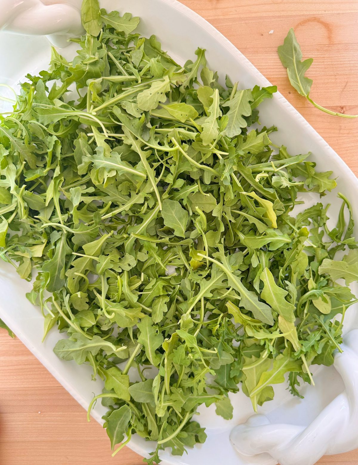 A white platter filled with fresh arugula leaves sits on a wooden surface.