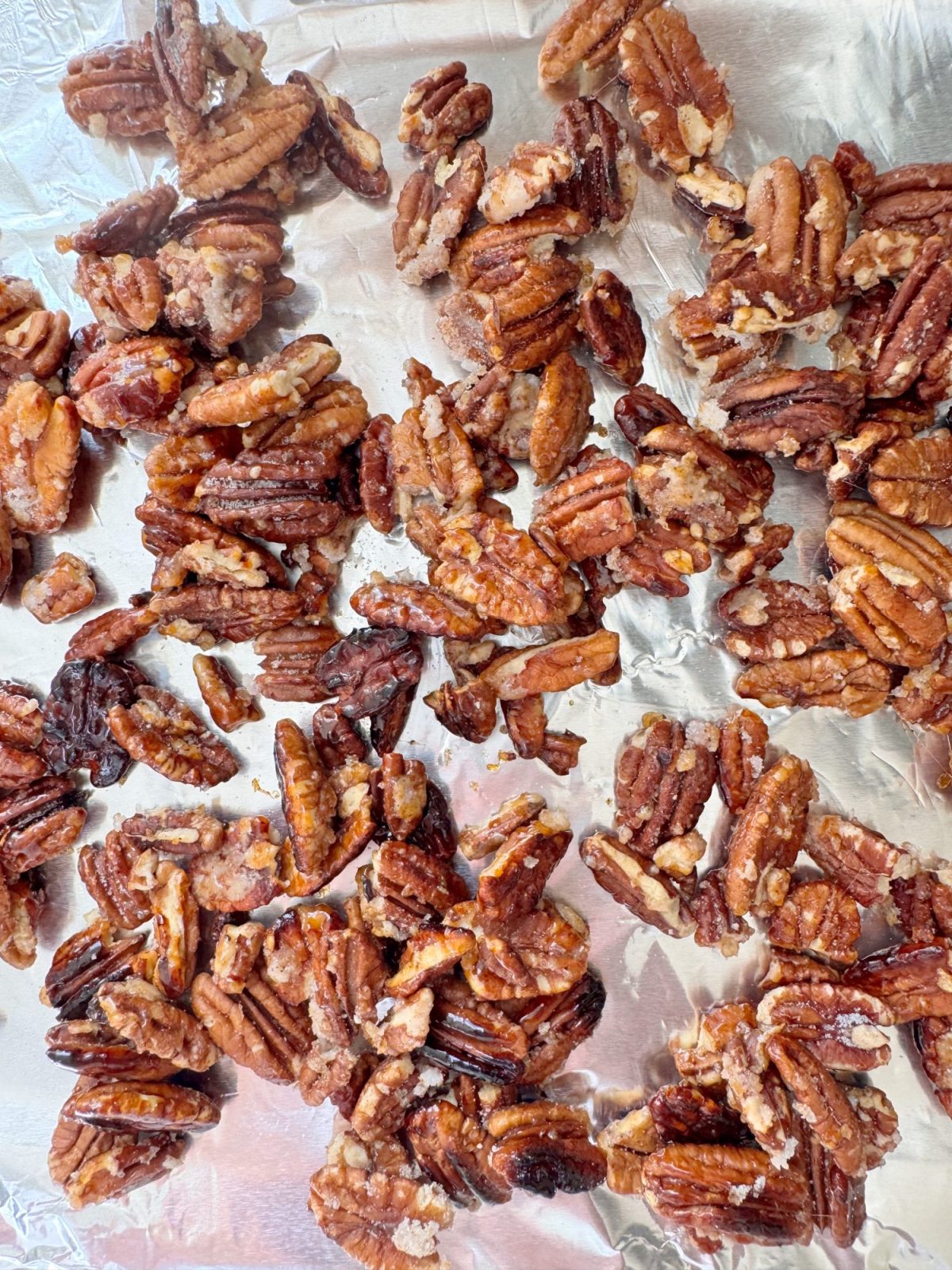 Pecan halves coated with sugar and spices are spread out on a sheet of aluminum foil.