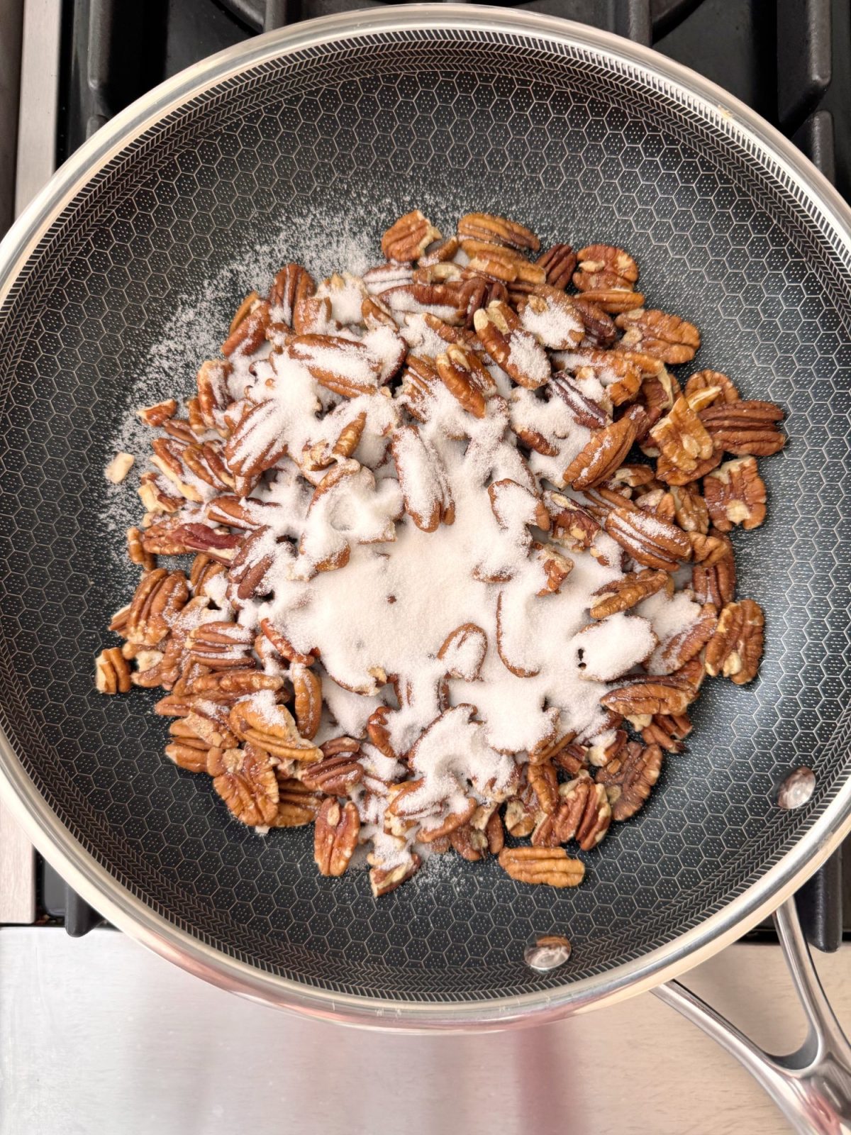 Pecan halves in a frying pan topped with granulated sugar, on a stove ready for cooking.