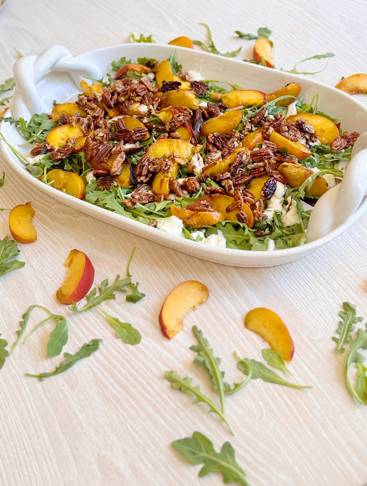 A white dish filled with arugula, sliced peaches, candied pecans, and cheese sits on a light wood table, with additional arugula and peach slices scattered around.