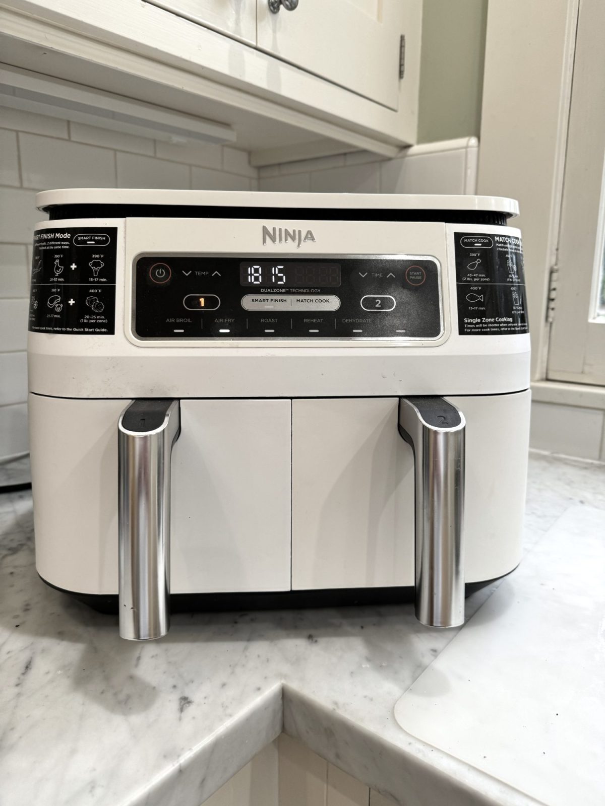 A white Ninja dual-basket air fryer with digital display and silver handles sits on a marble countertop in a kitchen.