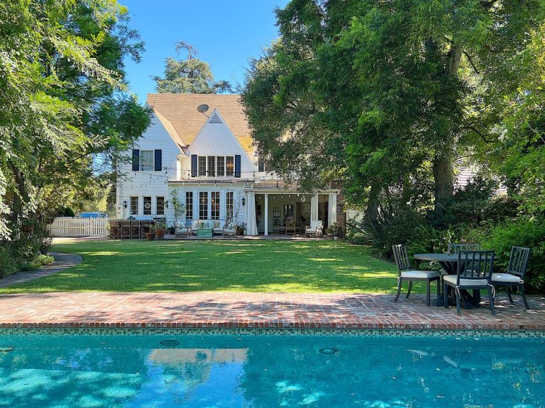 A white two-story house with a patio and outdoor seating faces a brick-edged swimming pool, surrounded by green lawn and trees&mdash;perfect for relaxing or streaming your favorite Amazon Prime shows outdoors.
