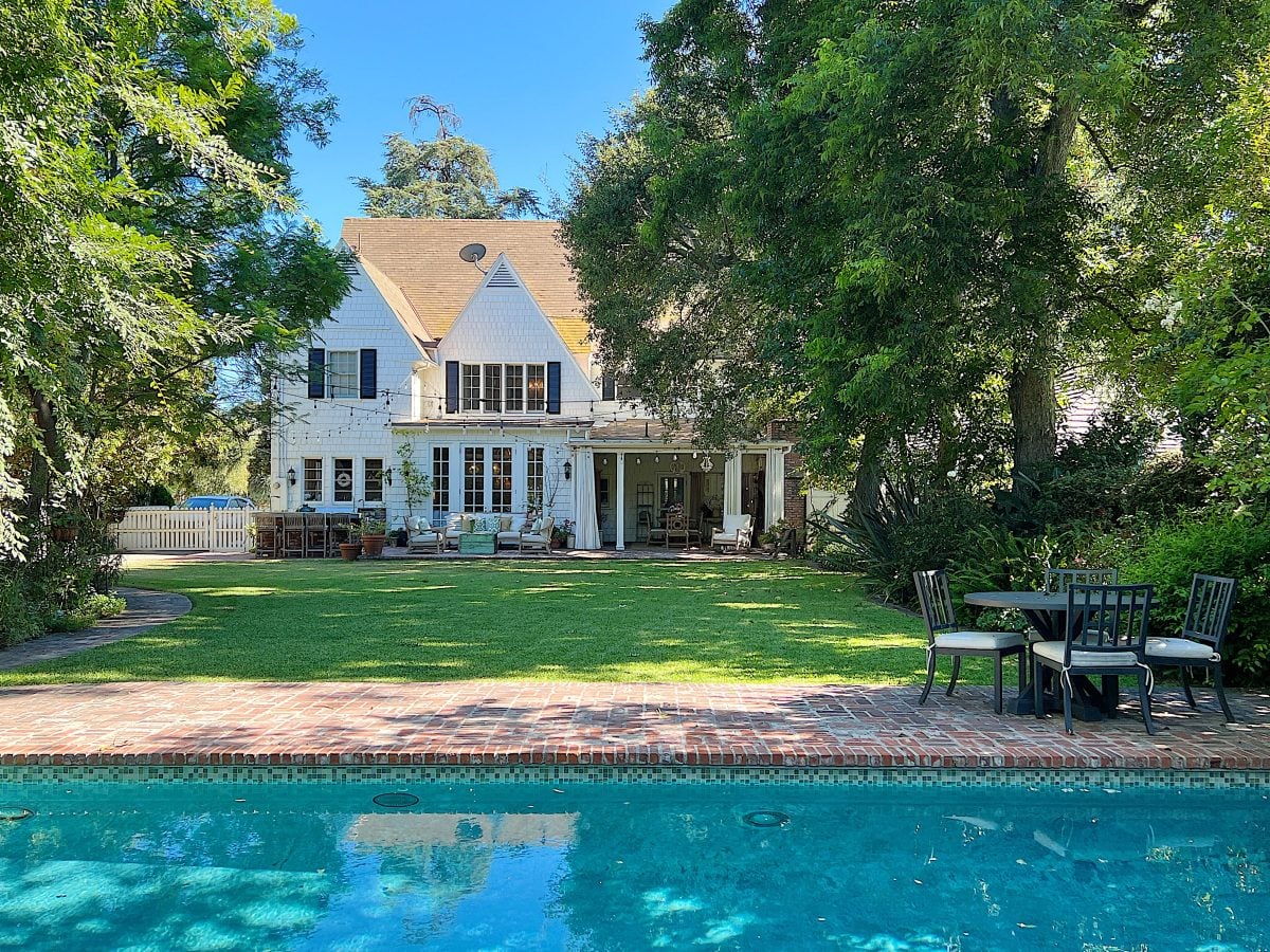 A white two-story house with a patio and outdoor seating faces a brick-edged swimming pool, surrounded by green lawn and trees—perfect for relaxing or streaming your favorite Amazon Prime shows outdoors.