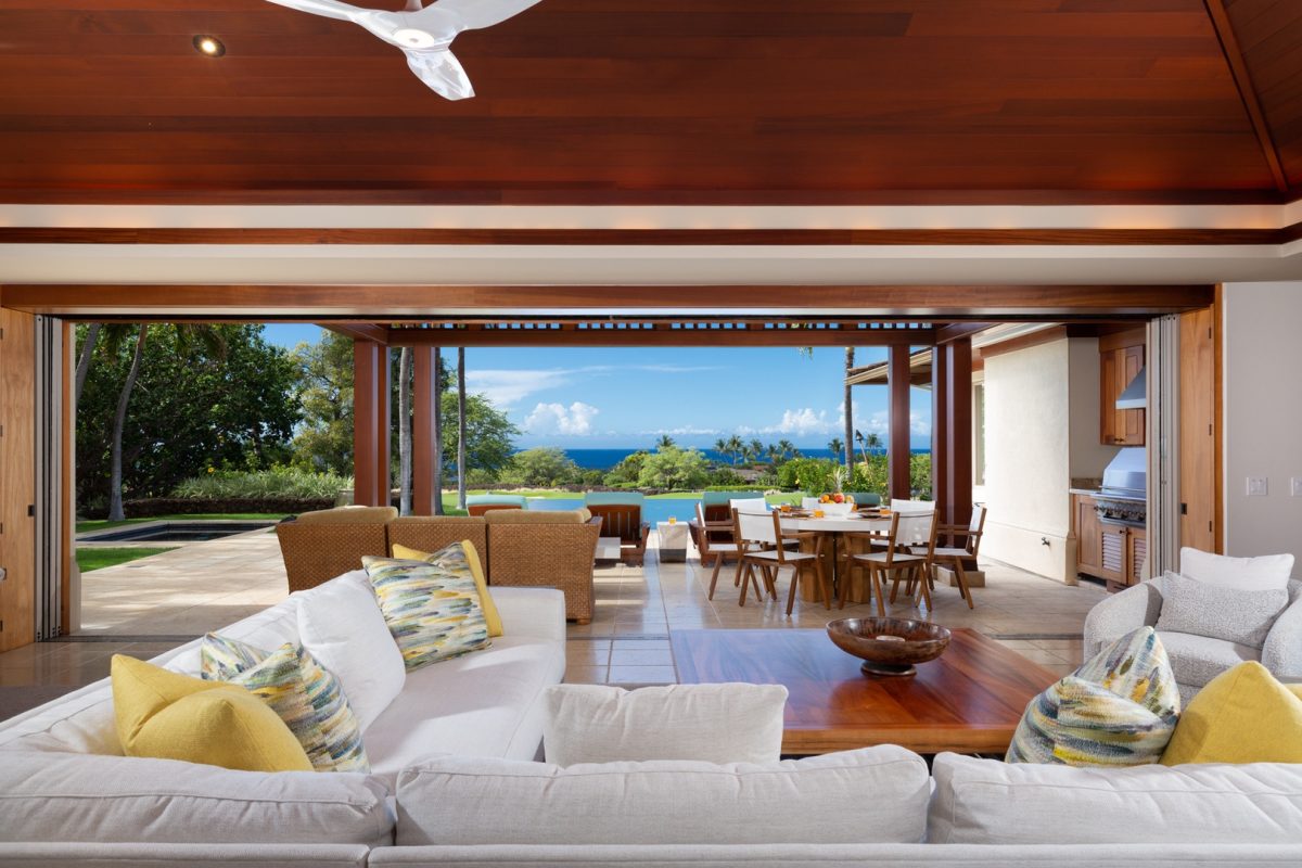 Spacious open-concept living and dining area with white sofas, wooden furniture, and large sliding doors overlooking a patio, pool, and ocean view.