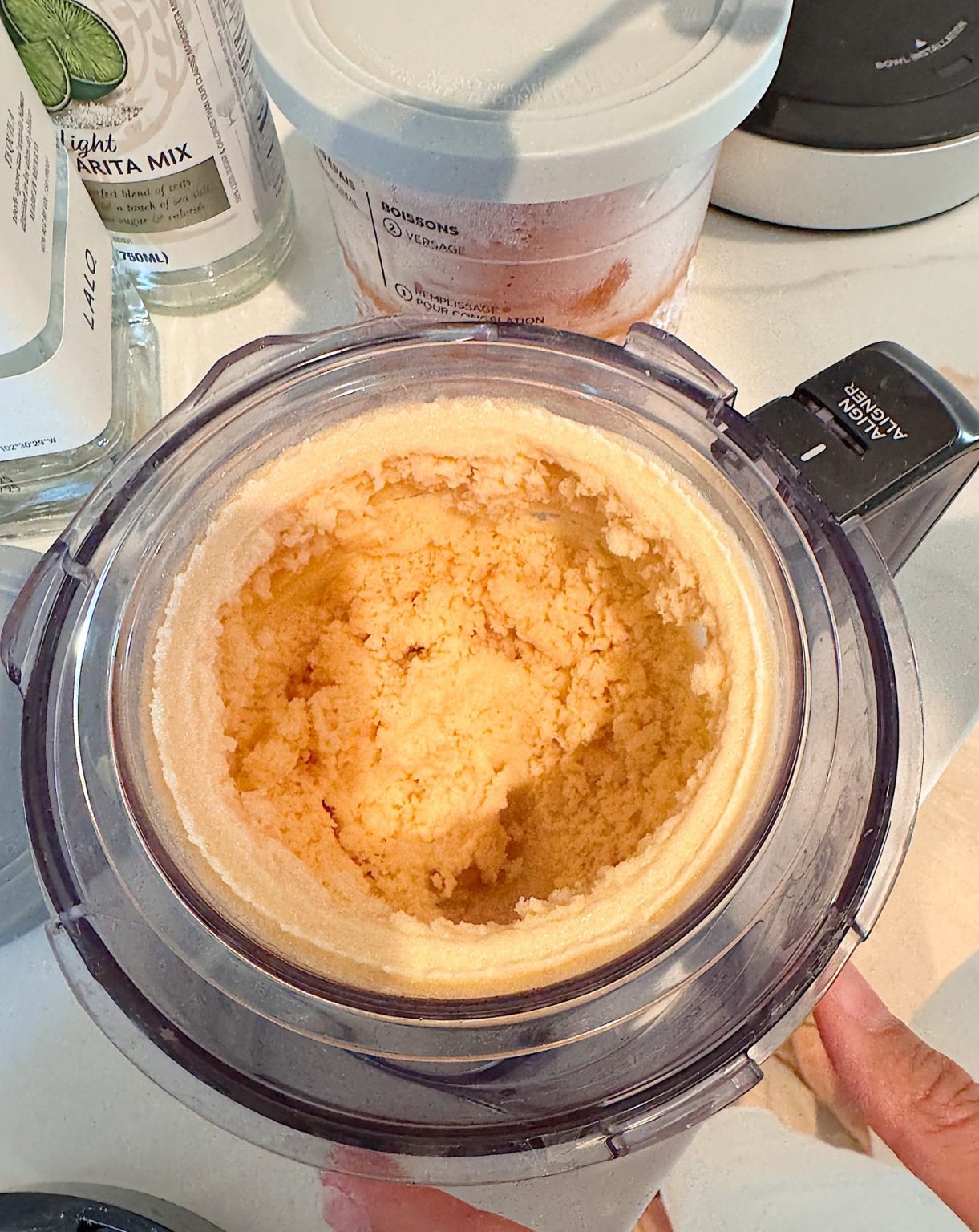 A close-up of a countertop ice cream maker containing partially churned ice cream, with ingredients and containers visible in the background.