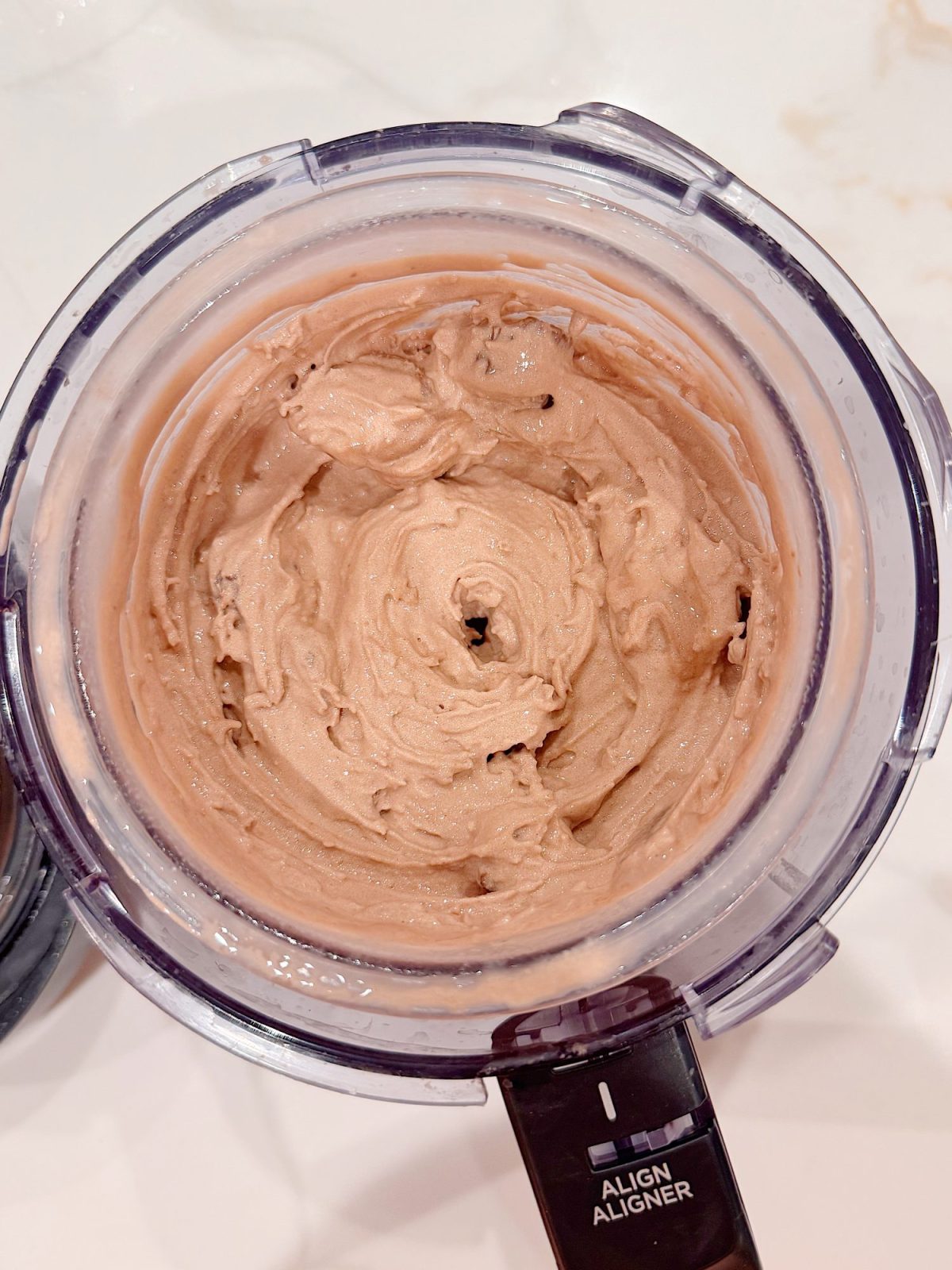 Chocolate ice cream mixture in a food processor, viewed from above, with smooth and creamy texture visible inside the bowl.