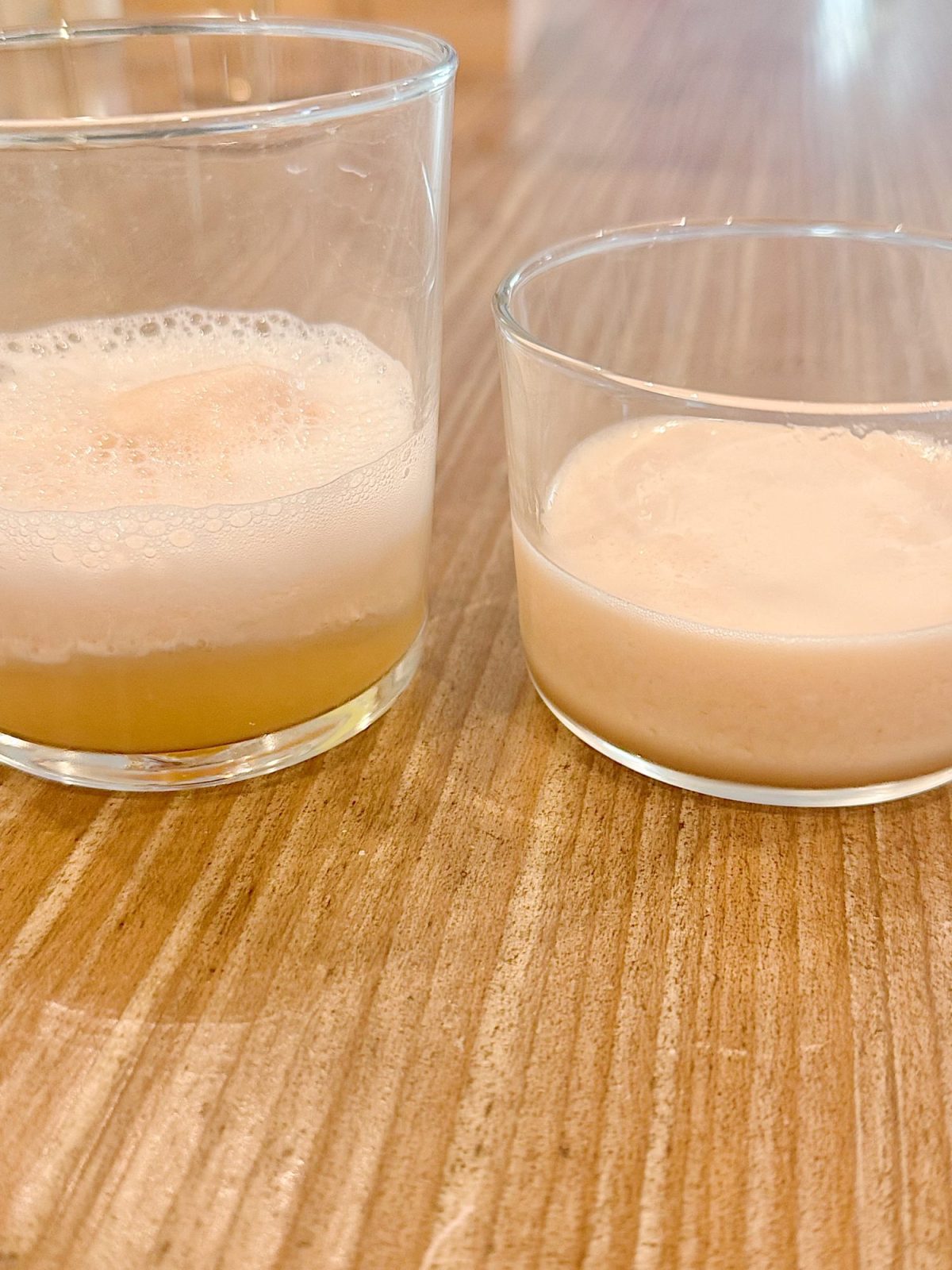 Two clear glasses with frothy beverages sit on a wooden surface; the left glass contains a yellow liquid, while the right glass contains a beige liquid.