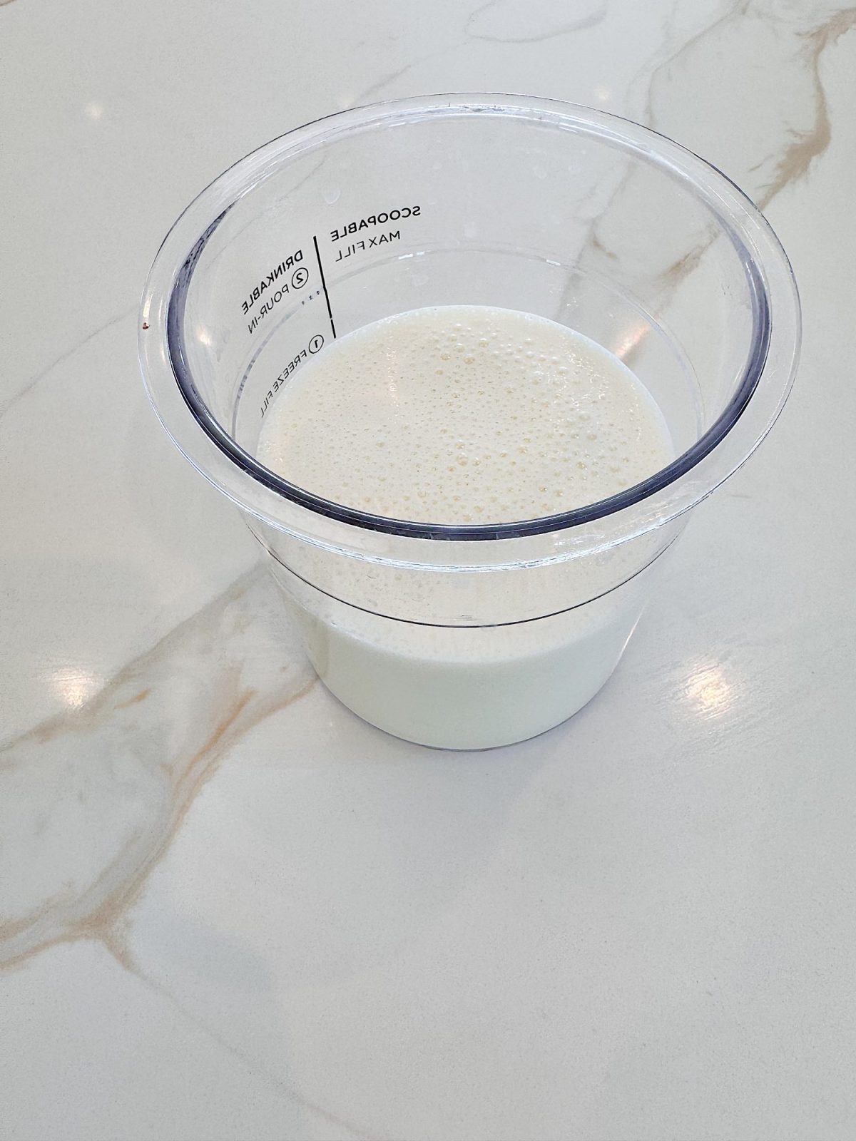 A clear plastic cup containing a portion of milk sits on a white, marbled surface.