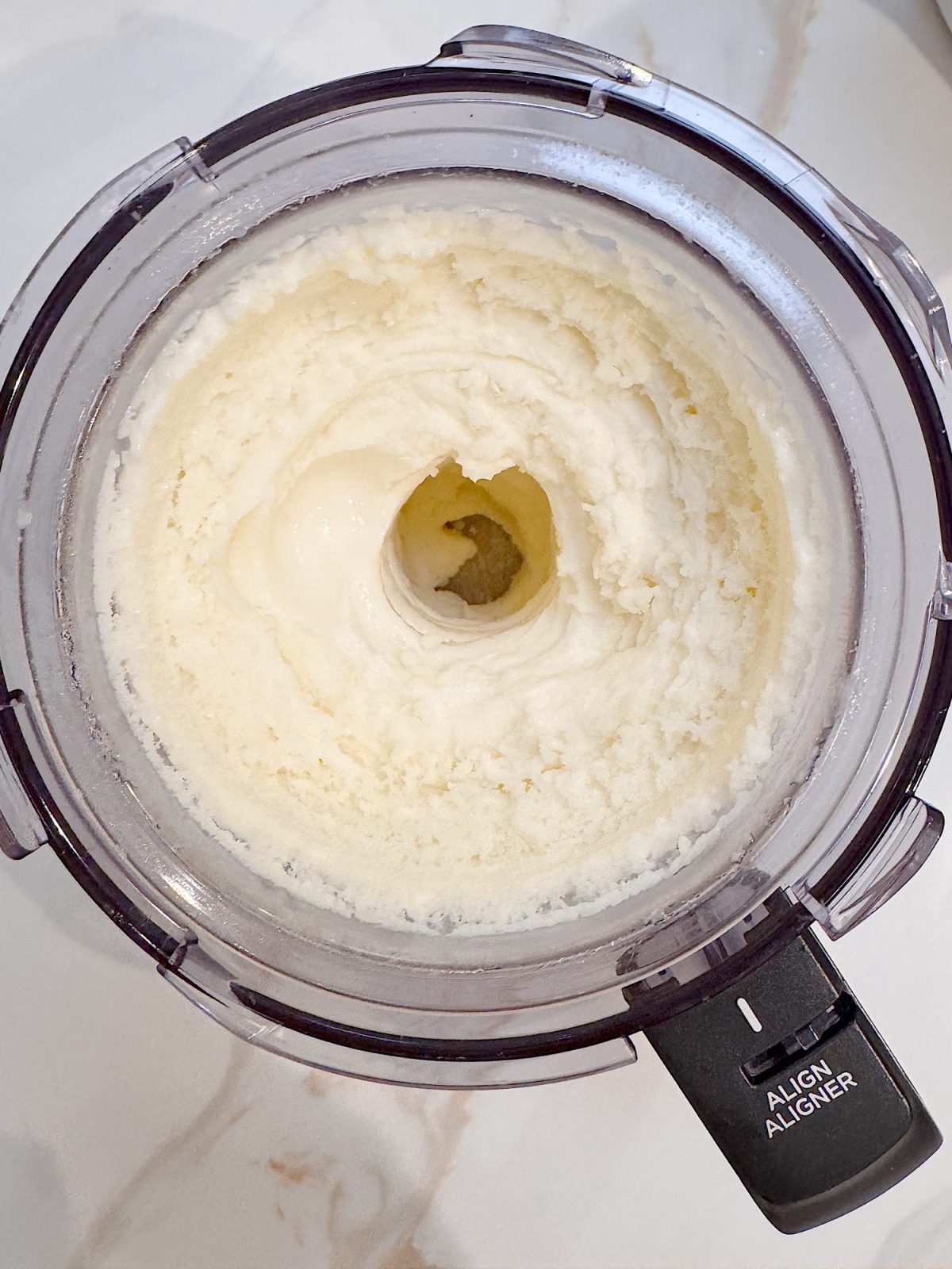 A food processor filled with blended, creamy white ice cream, shown from above on a white surface.