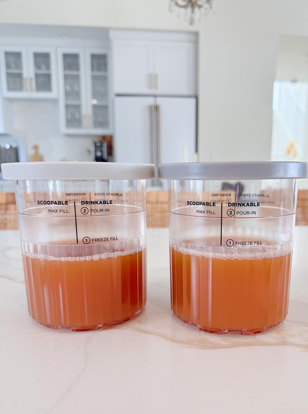 Two clear plastic containers with lids, partially filled with orange liquid, sit on a white kitchen counter; kitchen cabinets and appliances are visible in the background.