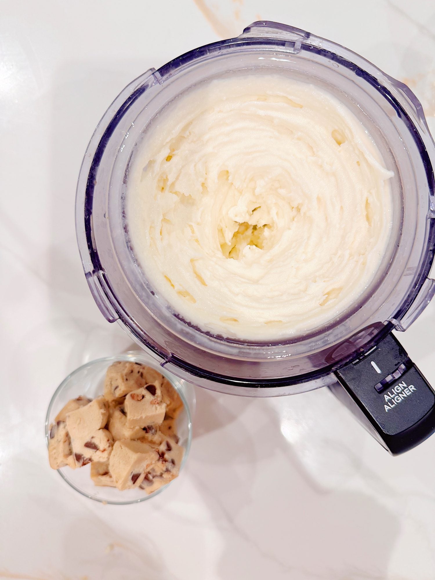 Overhead view of a food processor filled with blended ice cream next to a glass containing chunks of chocolate chip cookie dough on a white surface.