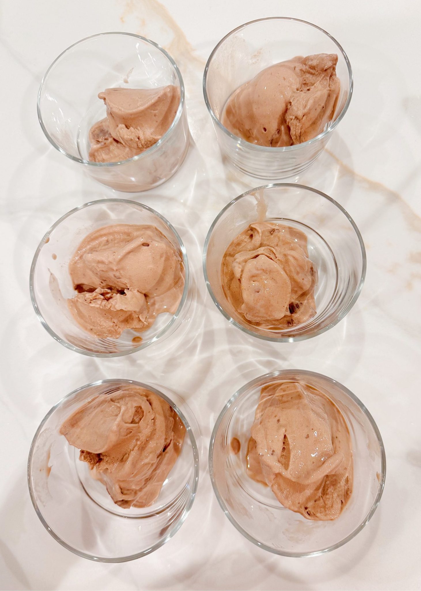 Six clear glasses each containing a scoop of chocolate ice cream are arranged in two rows of three on a white surface.