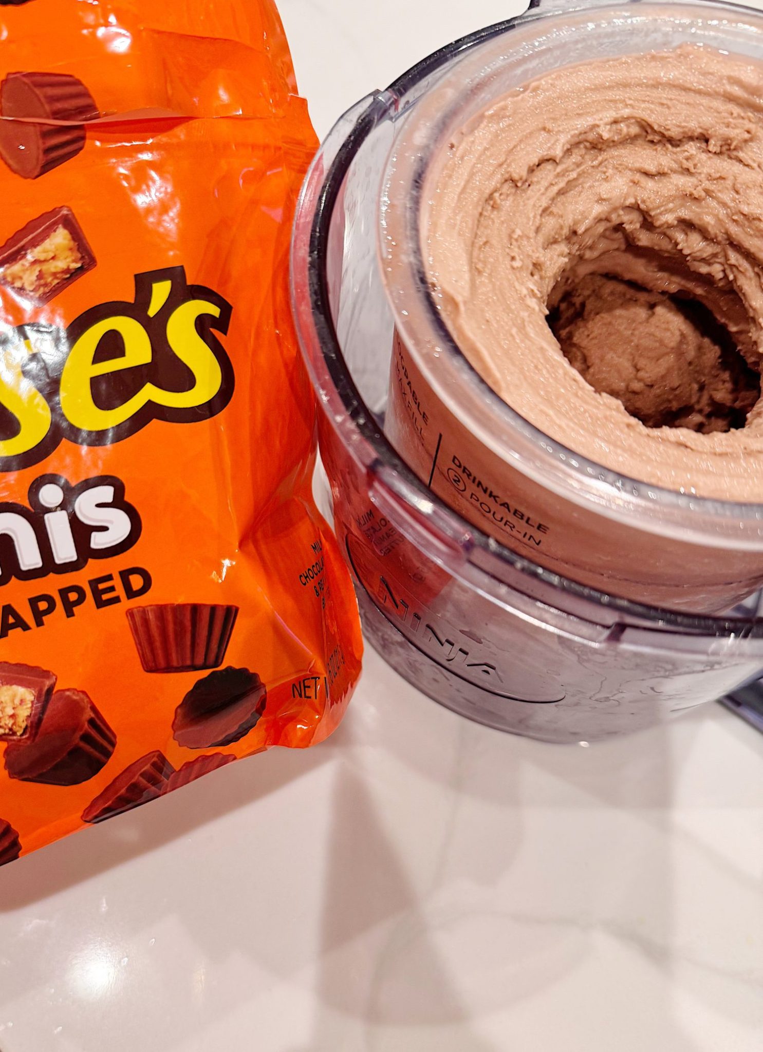 An open bag of Reese's Minis unwrapped chocolates next to a food processor containing chocolate mixture on a white surface.