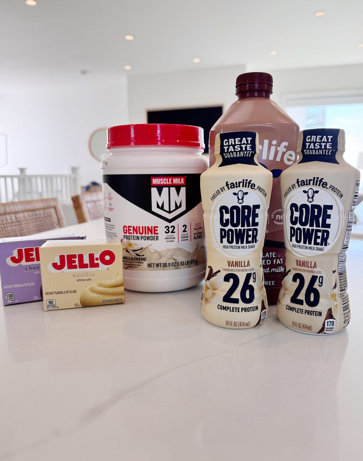 A variety of protein products, including Muscle Milk powder, Fairlife Core Power shakes, Fairlife chocolate milk, and two boxes of Jell-O, displayed on a kitchen counter.