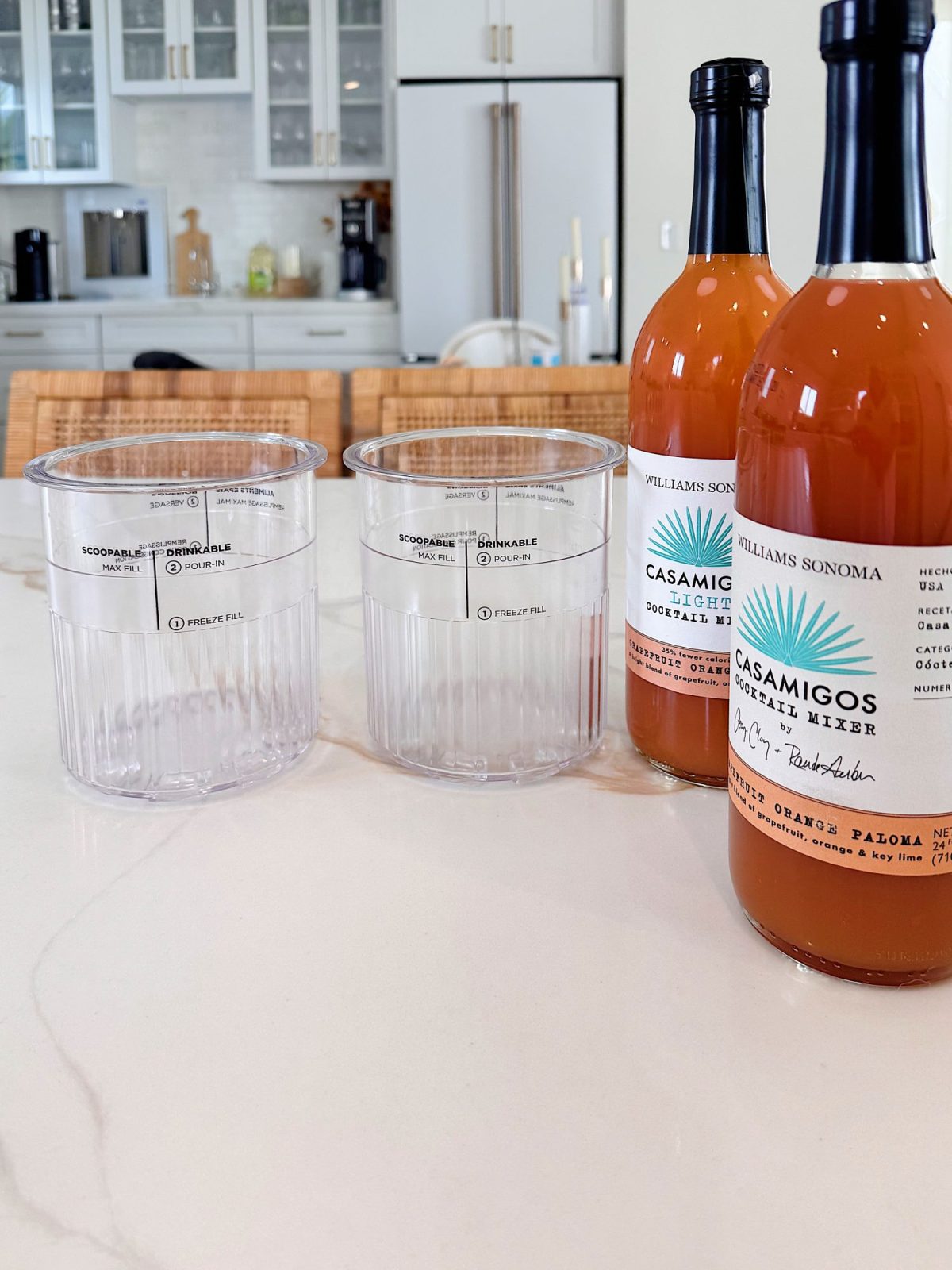 Two clear plastic cups and two bottles of Casamigos Cocktail Mixer are placed on a kitchen counter with a refrigerator and cabinets in the background.